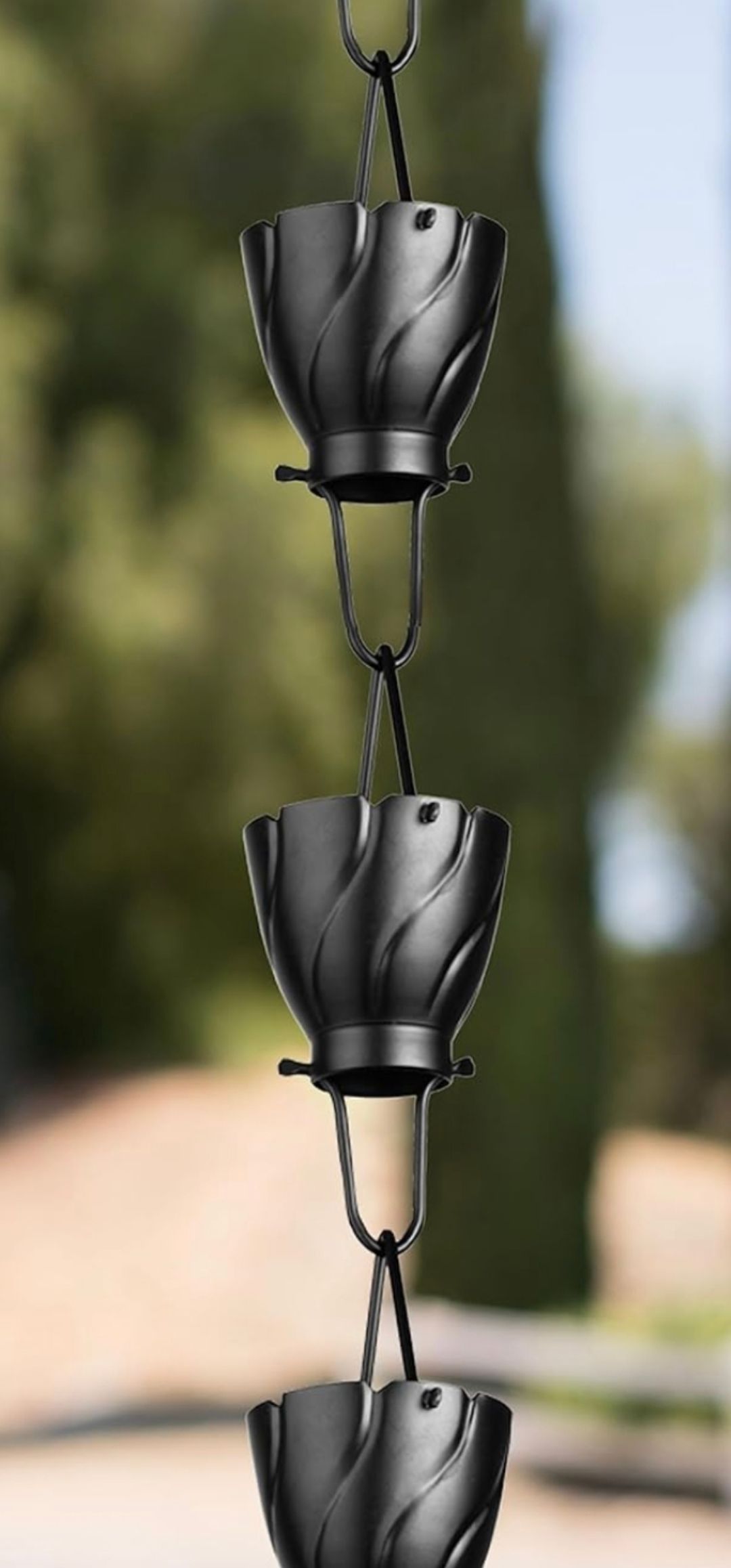 A black metal rain chain composed of repeating cup-shaped links with a spiral pattern, hanging against a blurred background.