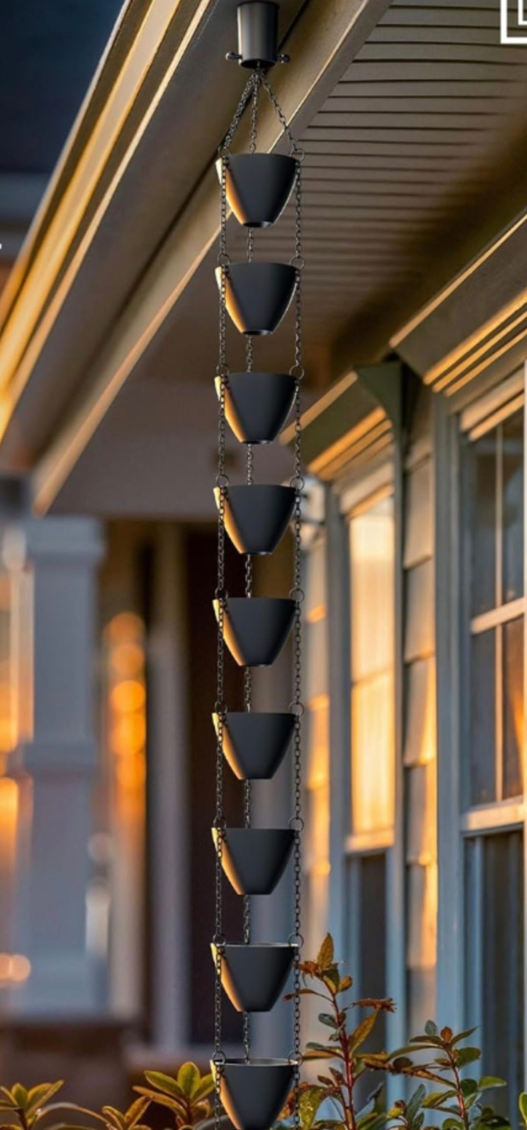 A black rain chain with connected metal cups hanging from a home's roof gutter.