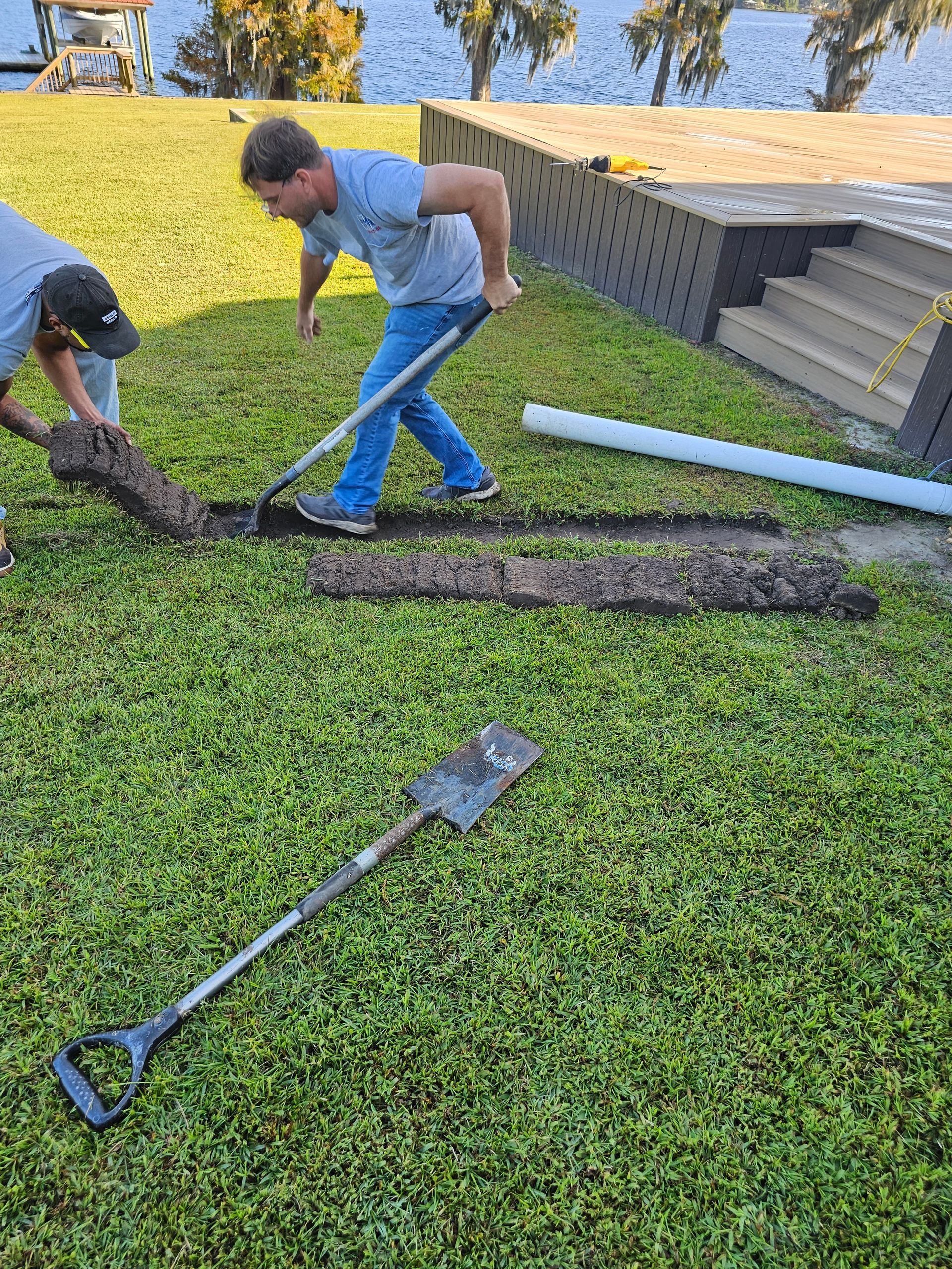 Two workers use shovels to dig a narrow trench through a green grassy lawn near a waterfront deck.