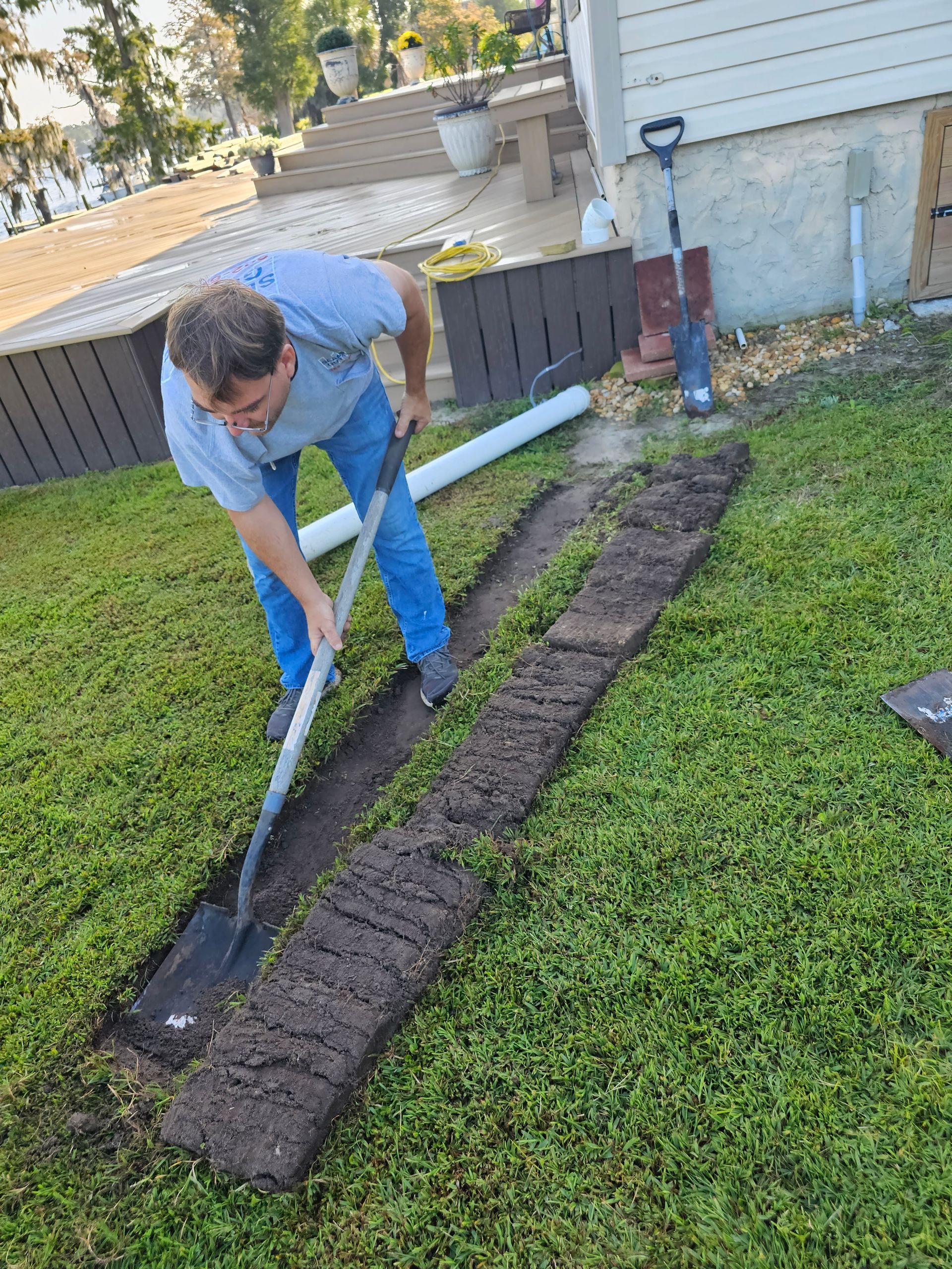 A person in a blue shirt and jeans uses a shovel to dig a trench in a grassy yard next to a white house.