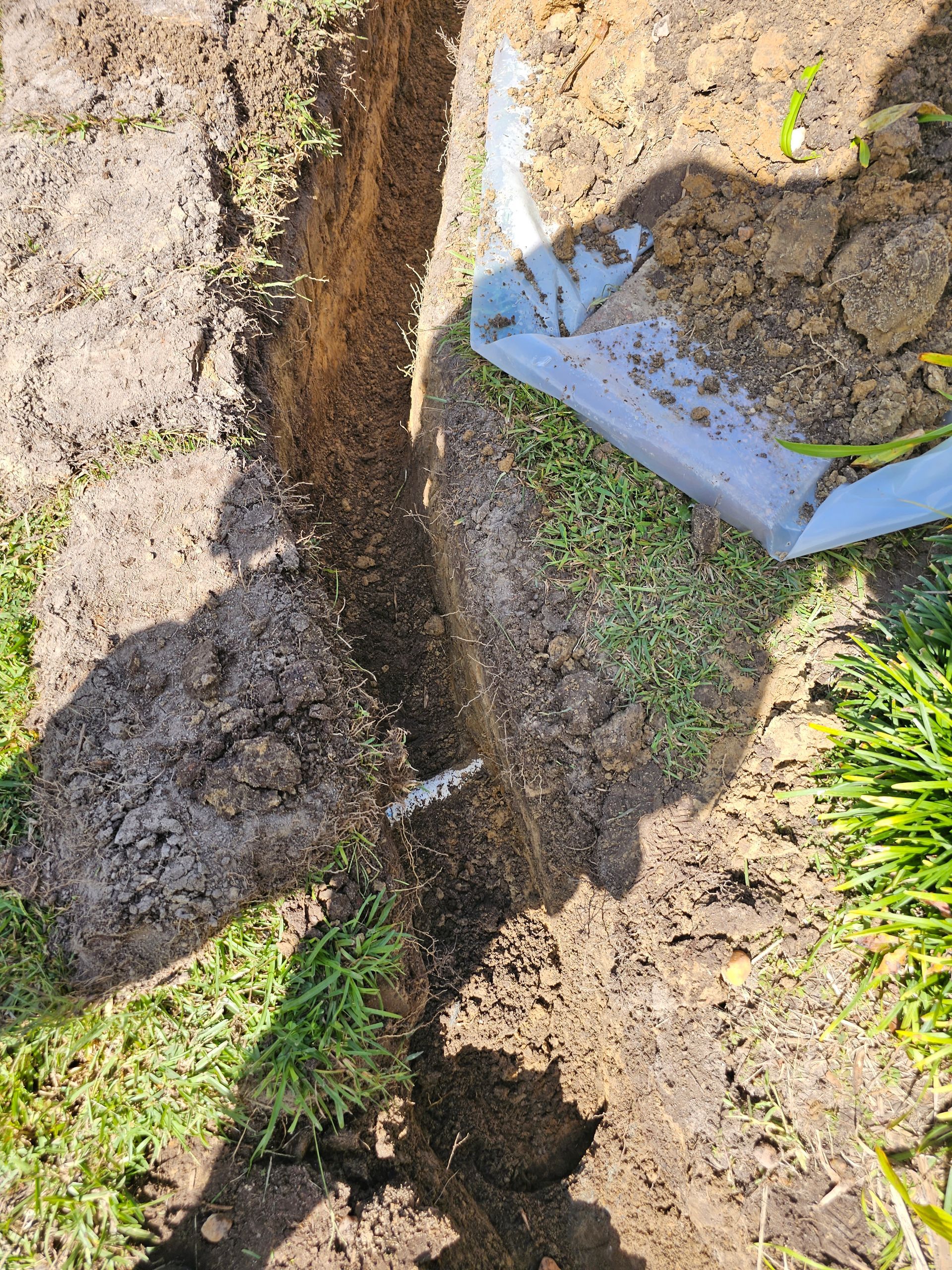 A narrow trench is dug into a grassy lawn, with a small section of white pipe visible inside and a blue plastic bag nearby.