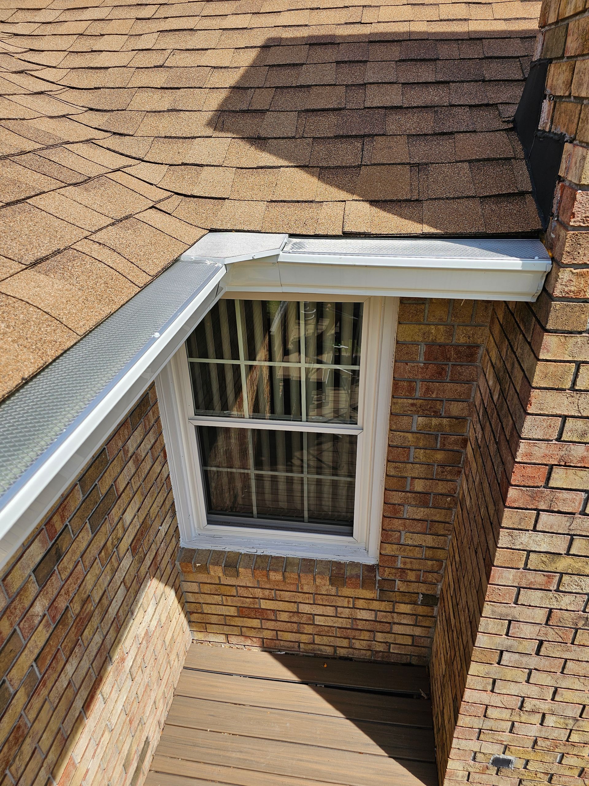 An outdoor view of a brick house corner, showing a shingled roof, a window, white gutters, and a wooden deck.