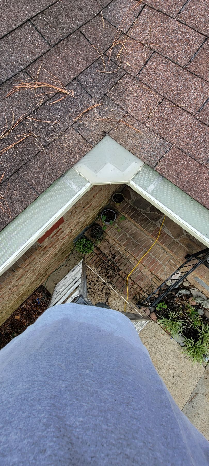 A high-angle view of a person standing on a ladder, looking down into a corner gutter filled with debris.