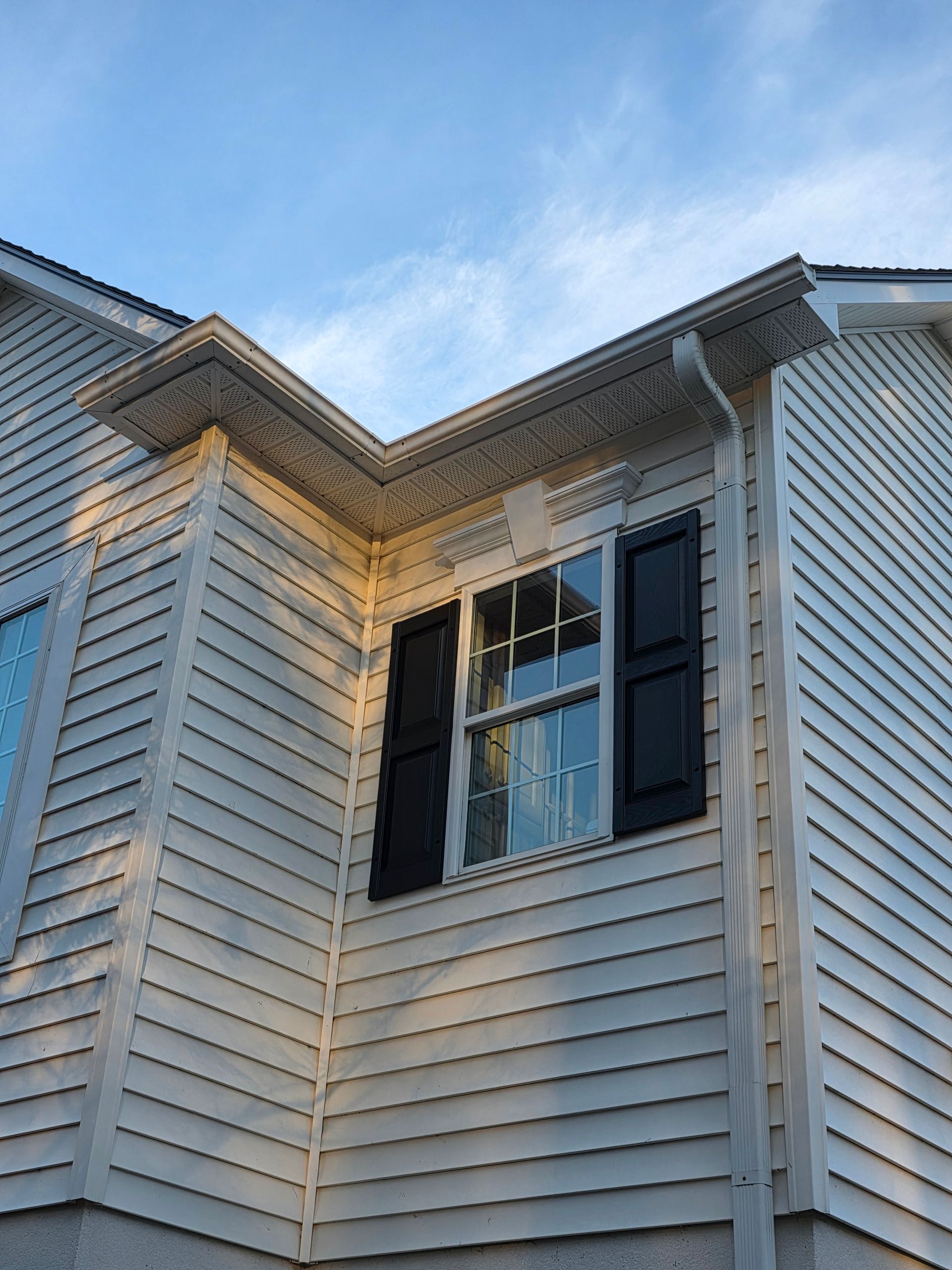 A close-up view of the corner of a light gray vinyl-sided house featuring a window with dark shutters and a downspout.