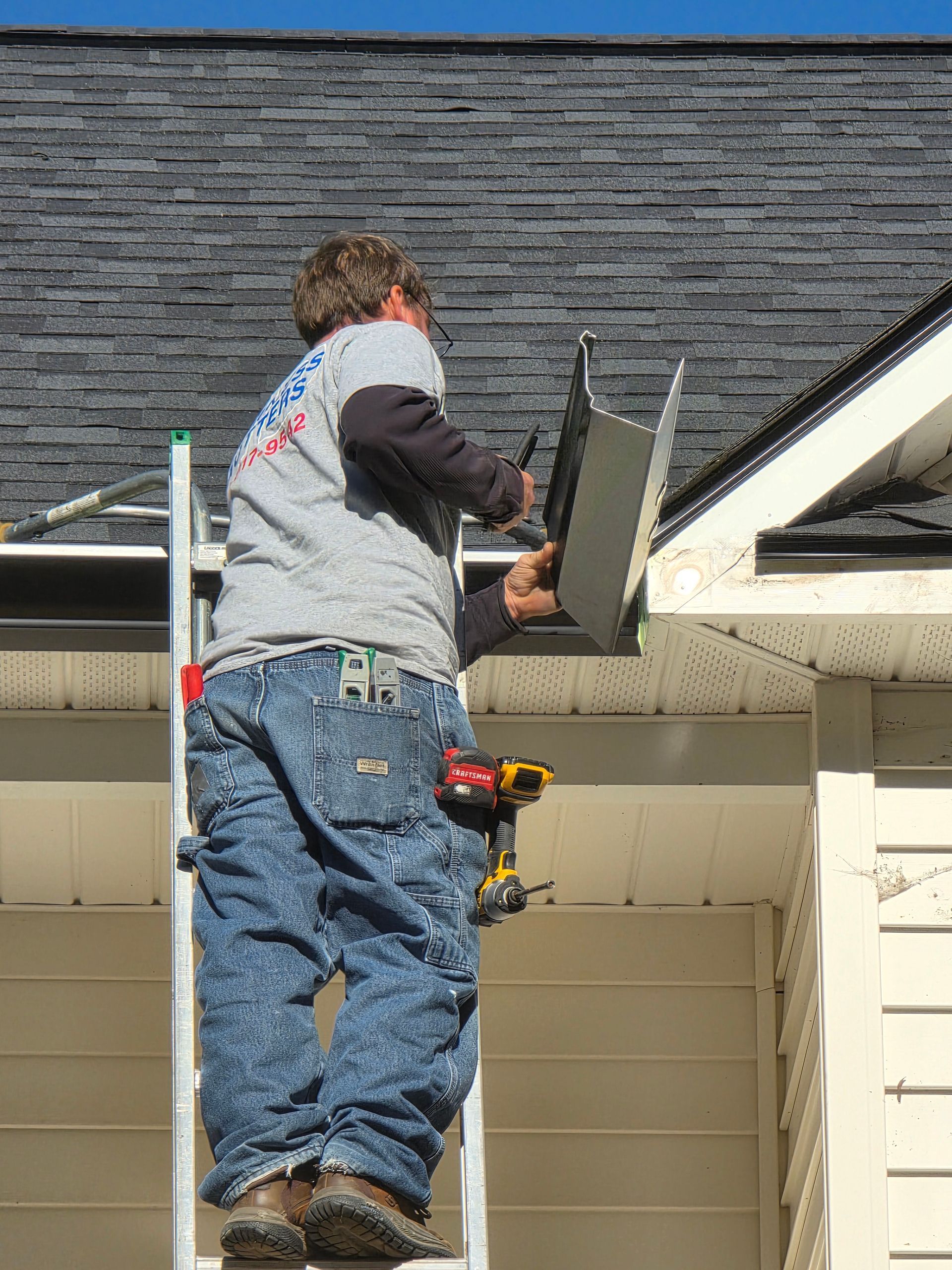 A person in a gray shirt and blue jeans stands on a ladder, installing a metal piece on the edge of a shingled roof.