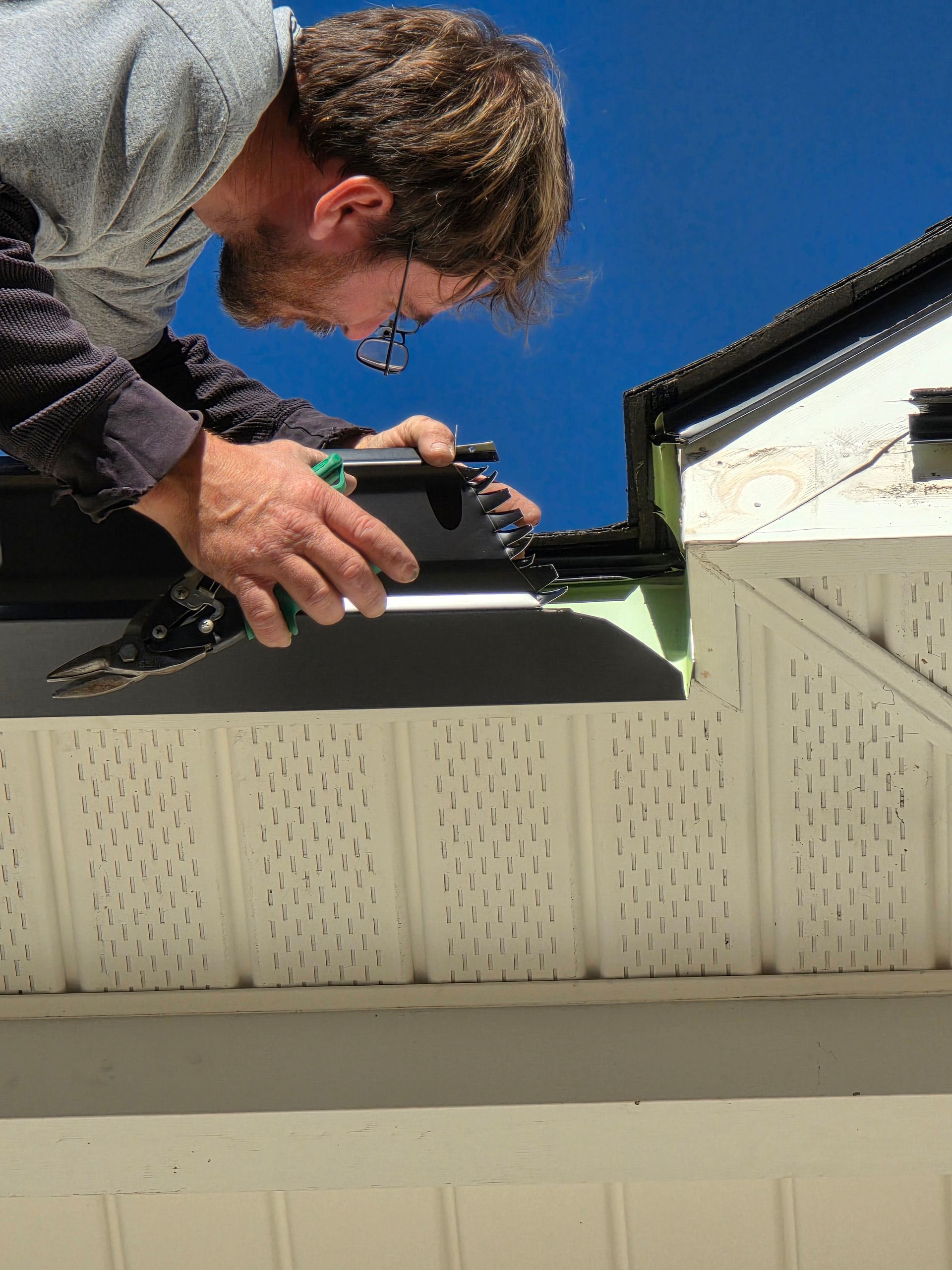 A worker attaches a dark metal gutter section to the edge of a roof against a bright blue sky.