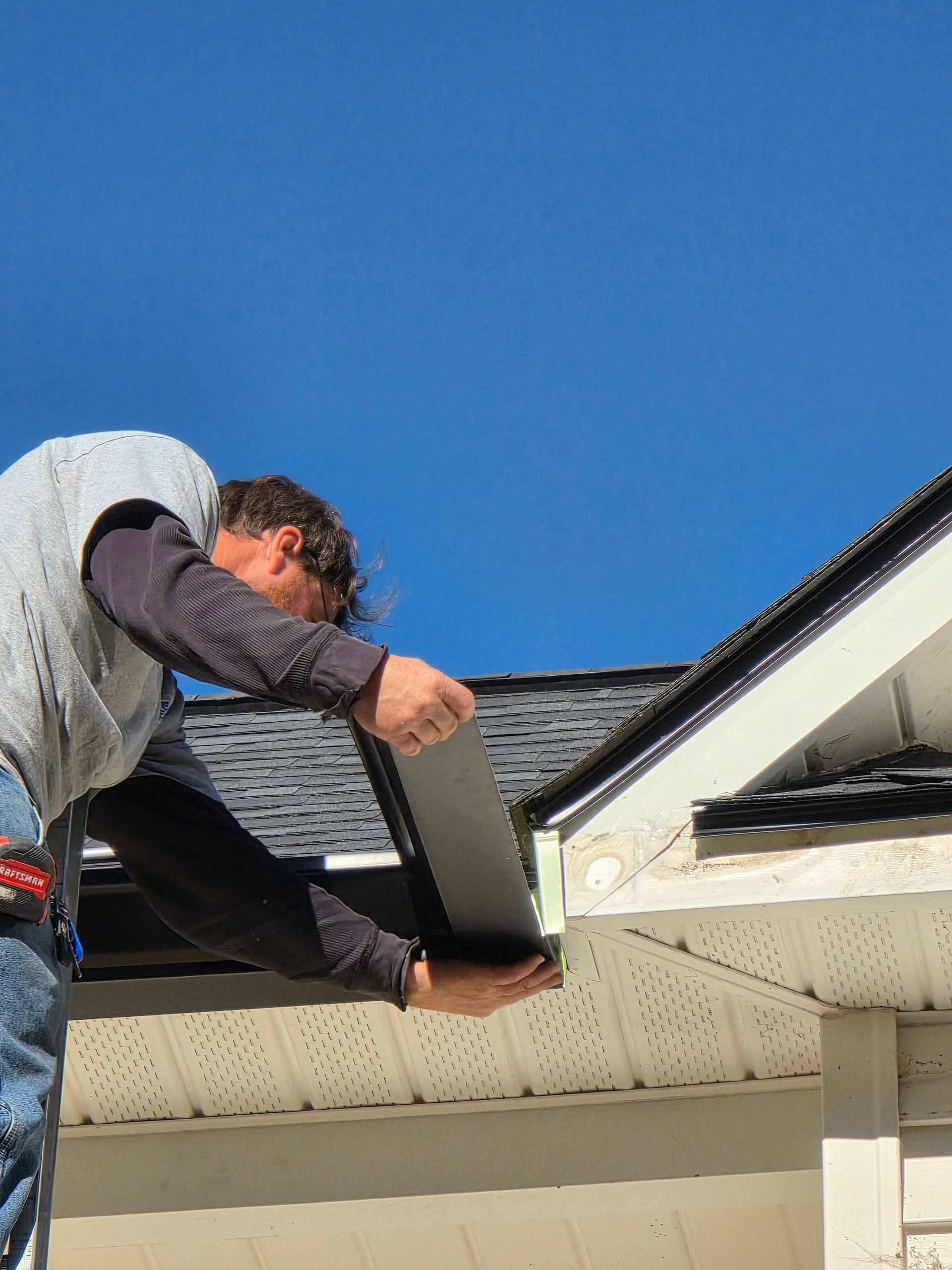 A person wearing a grey shirt installs dark metal flashing along the edge of a shingled roof against a clear blue sky.