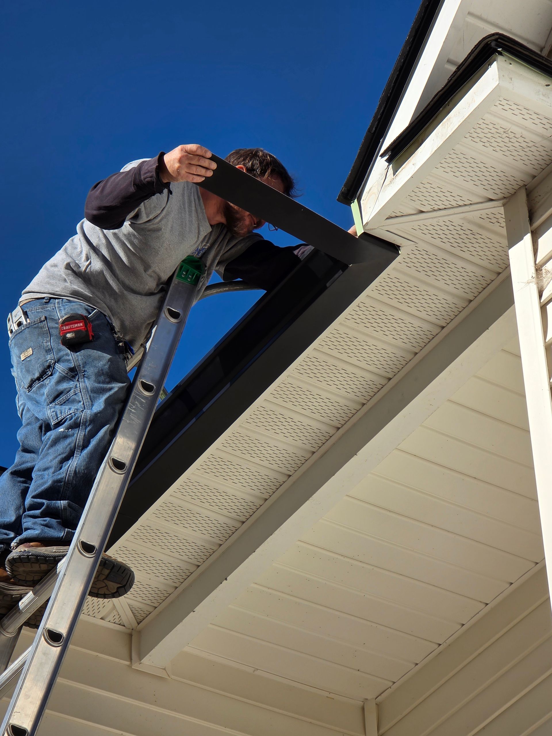 A worker standing on a ladder installs a black gutter along the edge of a house roof under a clear blue sky.