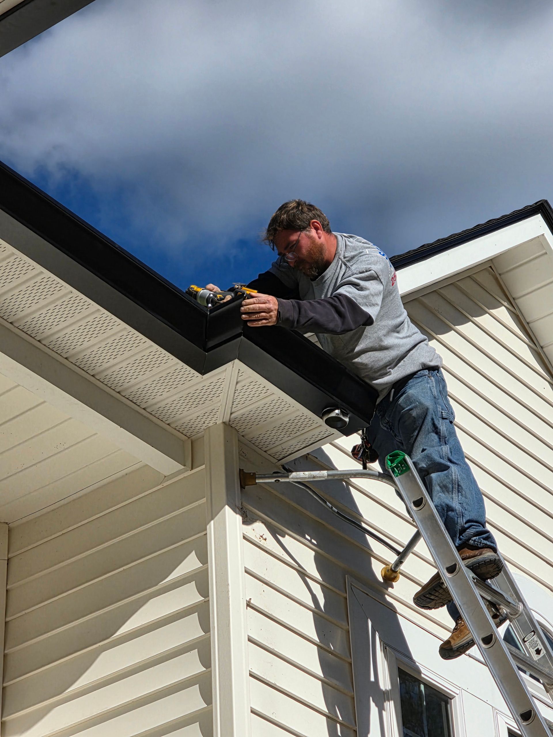 A person on a ladder repairs the black gutter and white soffit of a light-colored, siding-covered house under a blue sky.