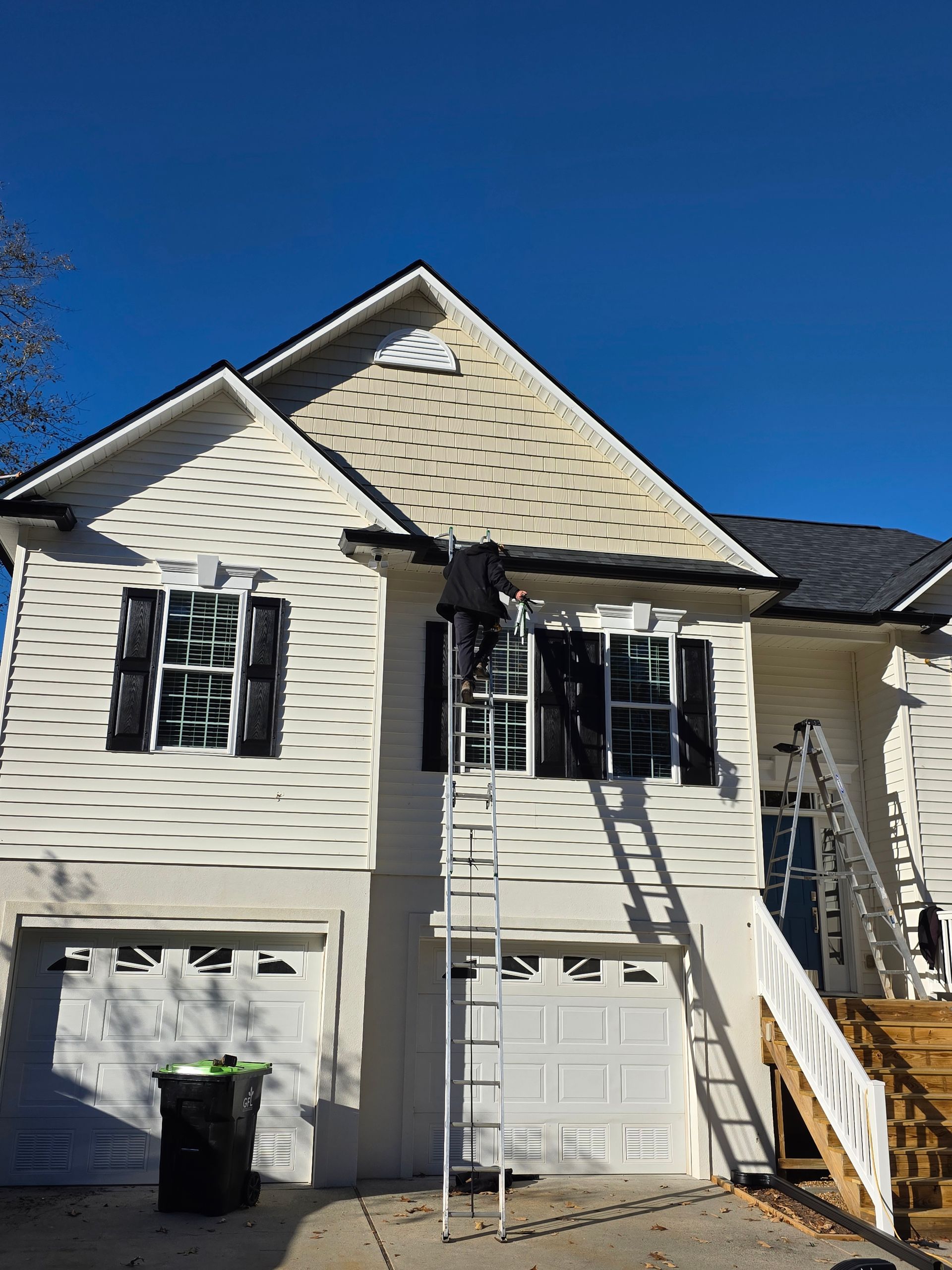 A person on a ladder repairs or cleans the exterior trim above a second-story window on a two-story beige house.