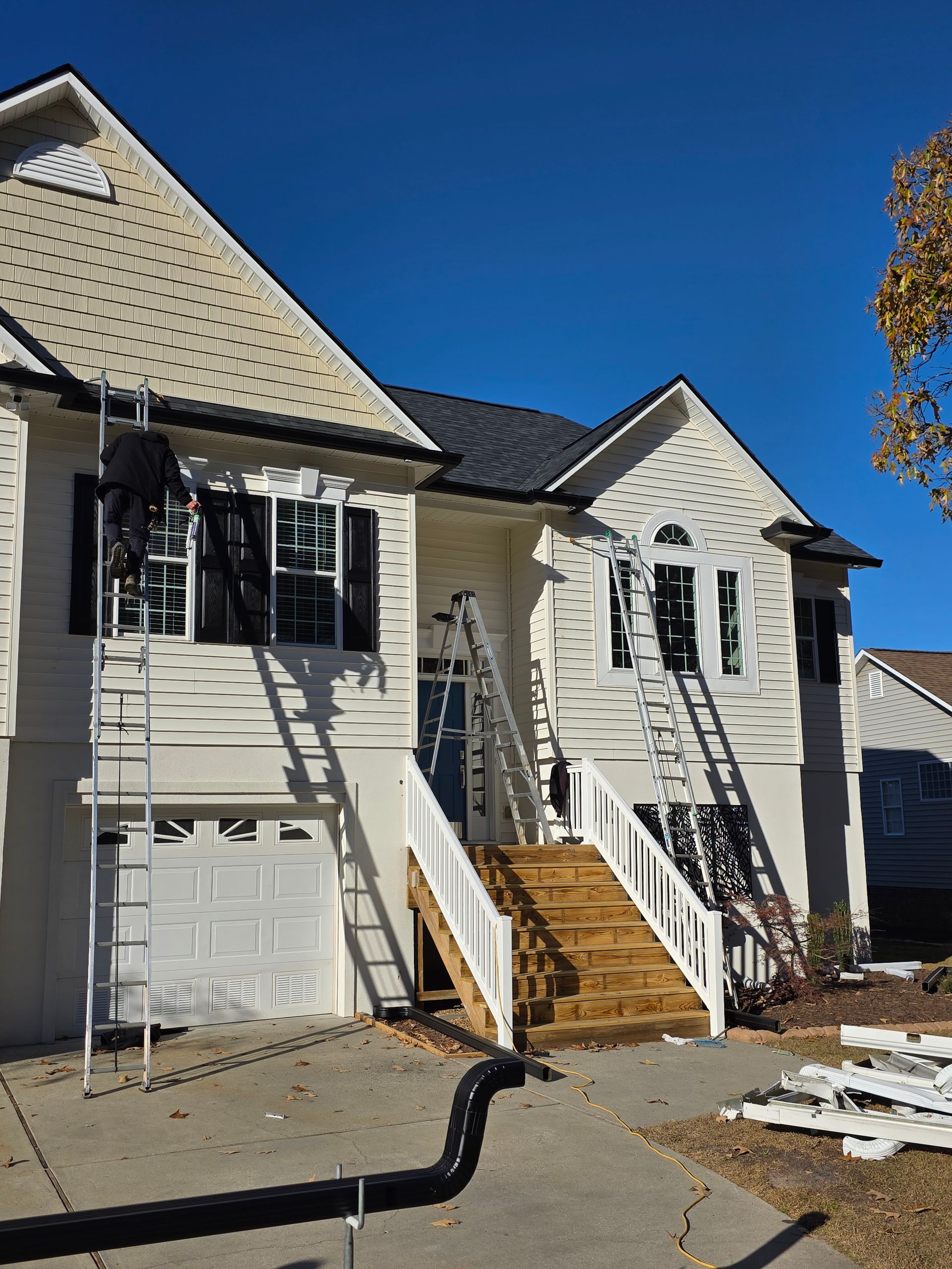 Workers use ladders to perform exterior home maintenance on a light-colored, multi-story house under a clear blue sky.