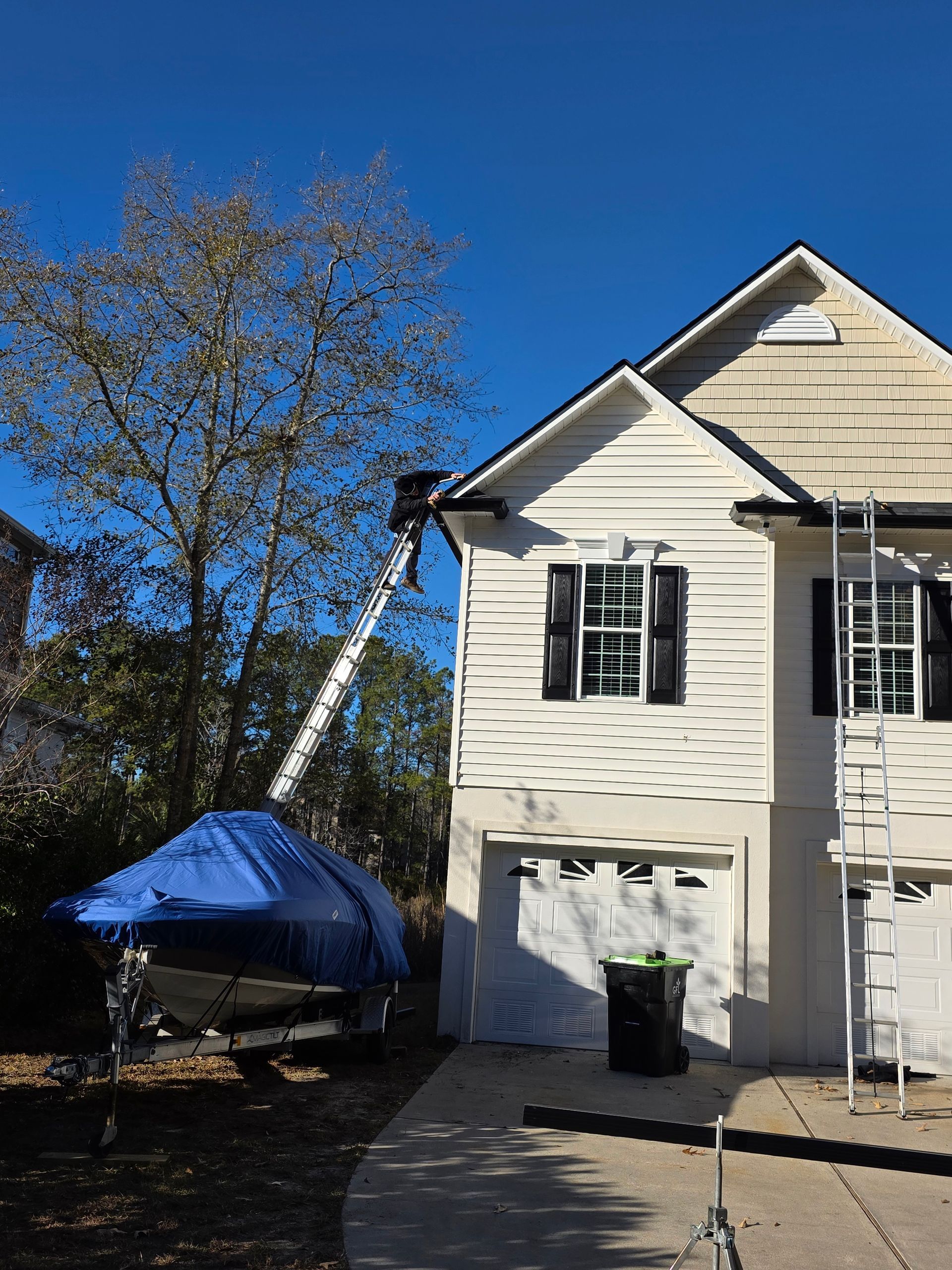 A multi-story house with cream siding, black shutters, and an extension ladder leaning against the roof near a boat.