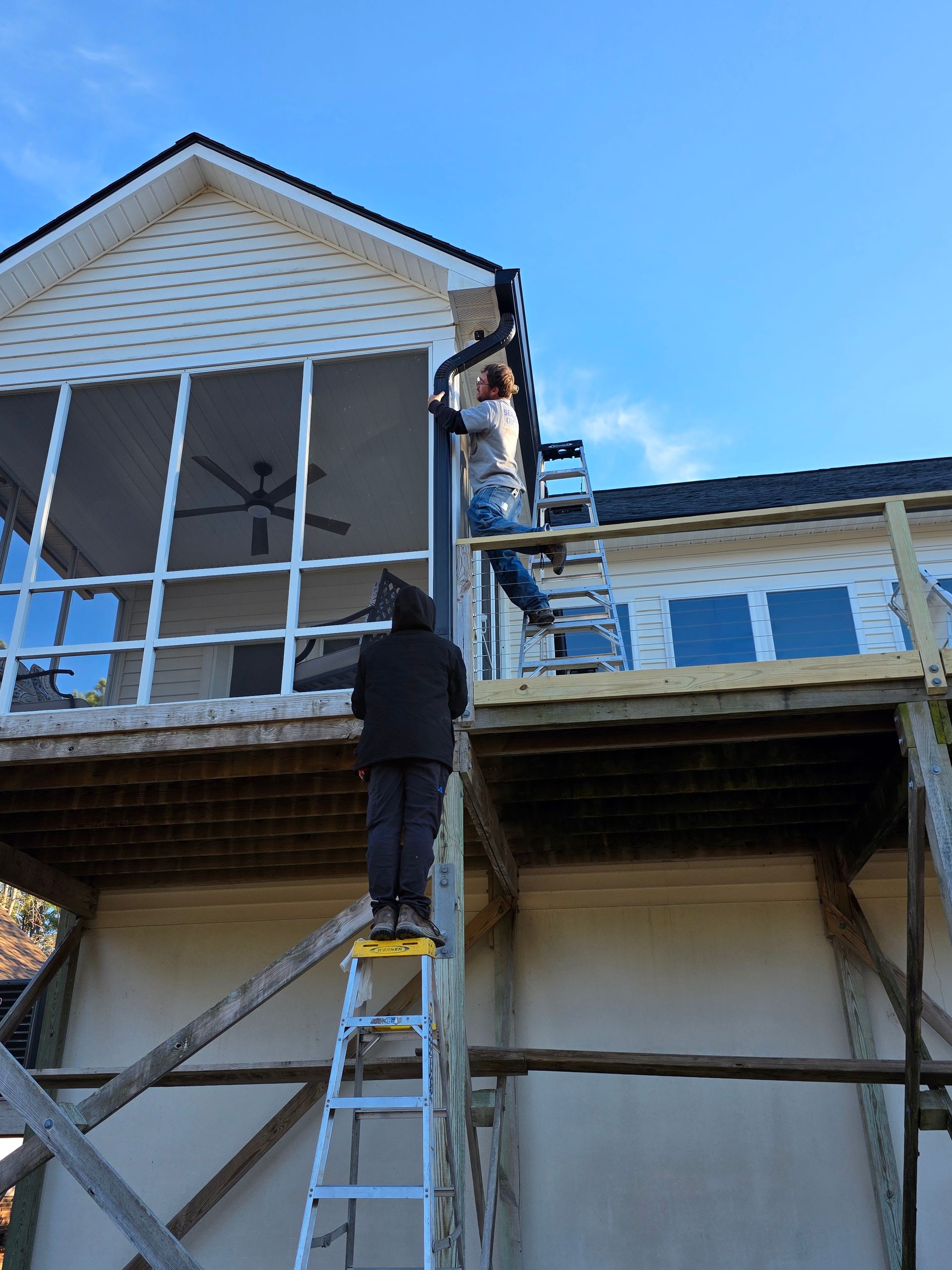 Two people work on a house exterior, using ladders to install gutters on a screened-in porch and deck under a blue sky.