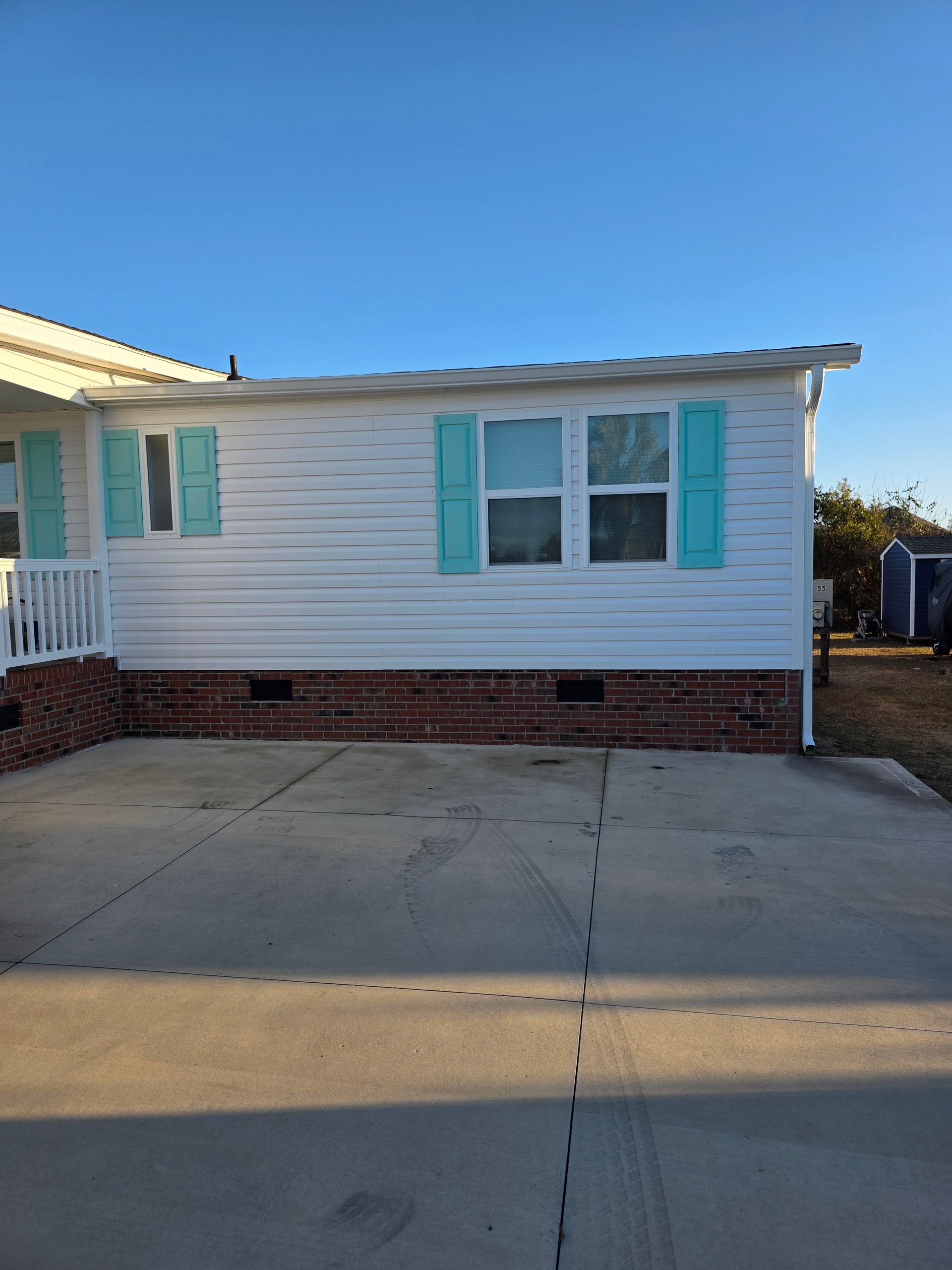 A white manufactured home with teal shutters and a brick foundation base, viewed from a concrete driveway.