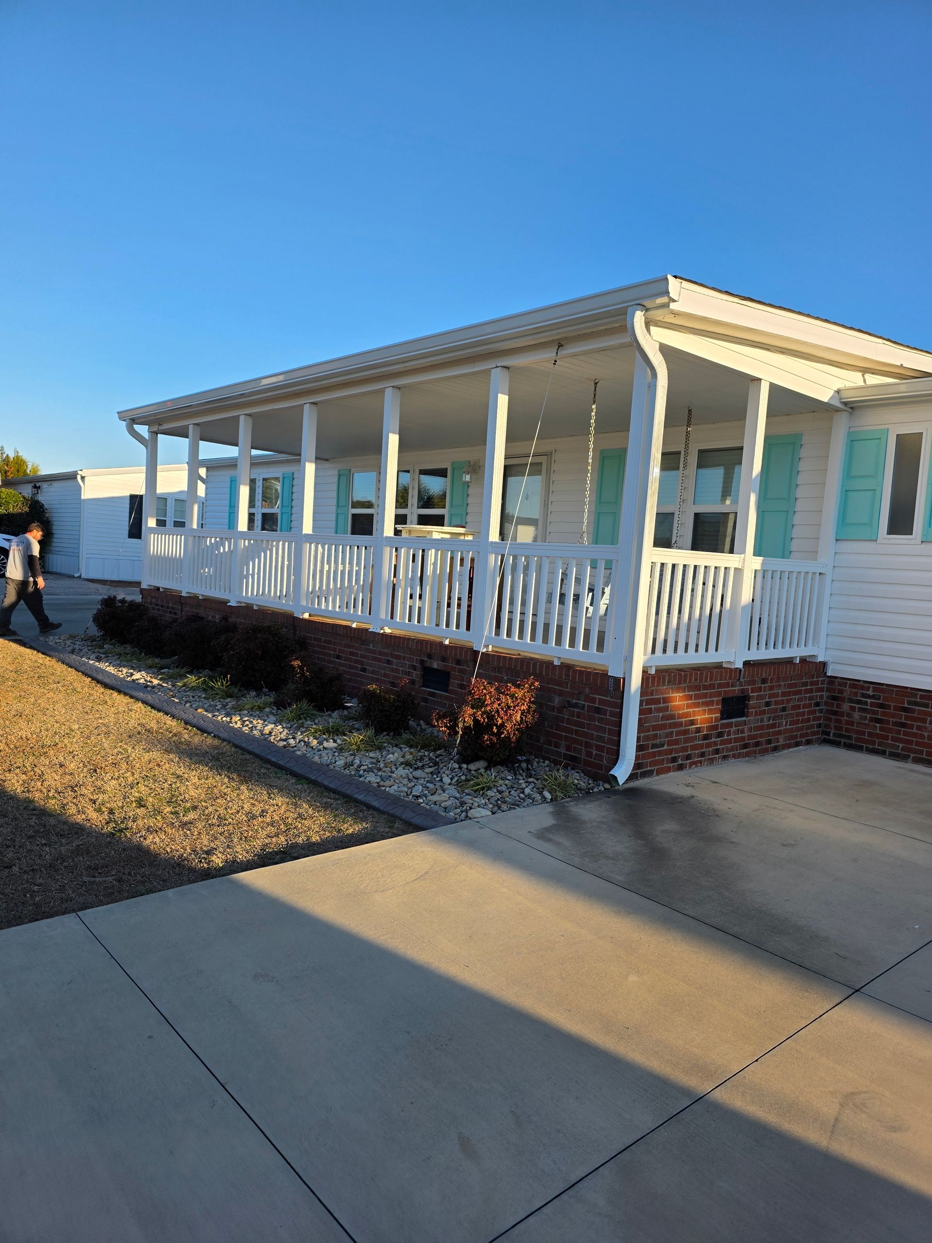 White mobile home featuring a large covered front porch, teal shutters, and a brick-patterned foundation under a clear sky.