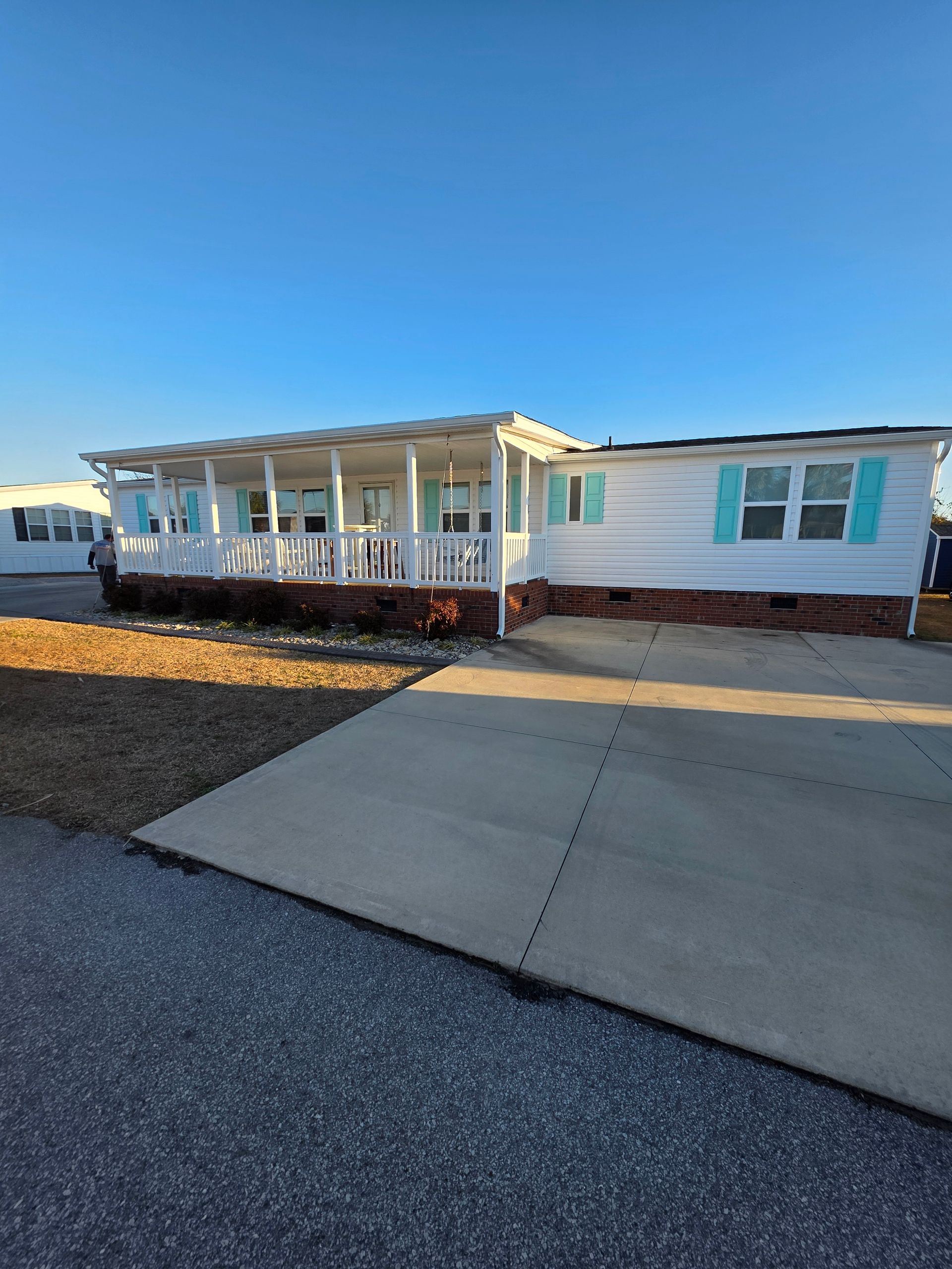 A white manufactured home with a large front porch, teal shutters, and a concrete driveway under a clear blue sky.