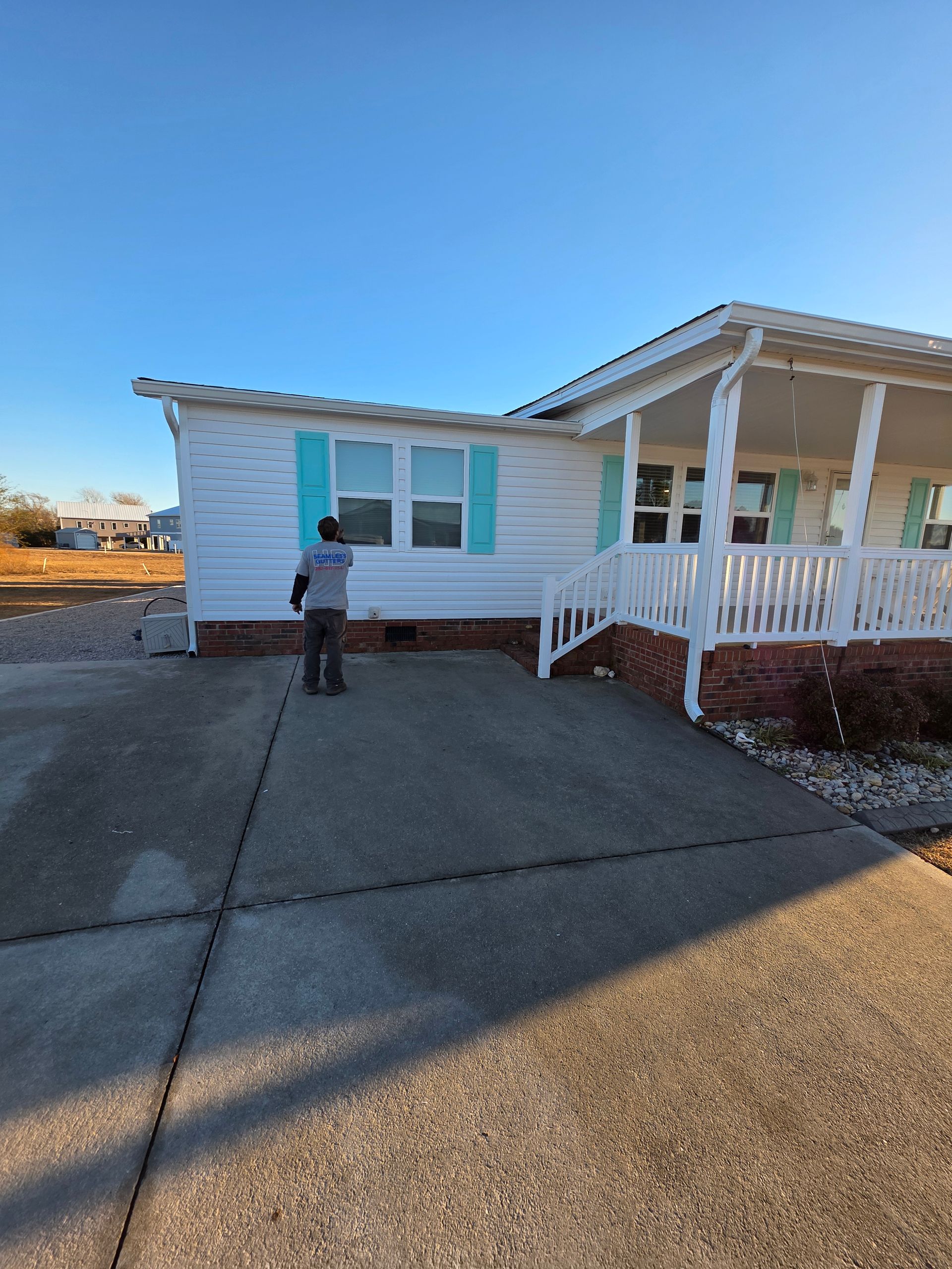 A person paints the window shutters of a white house with a porch under a clear blue sky.