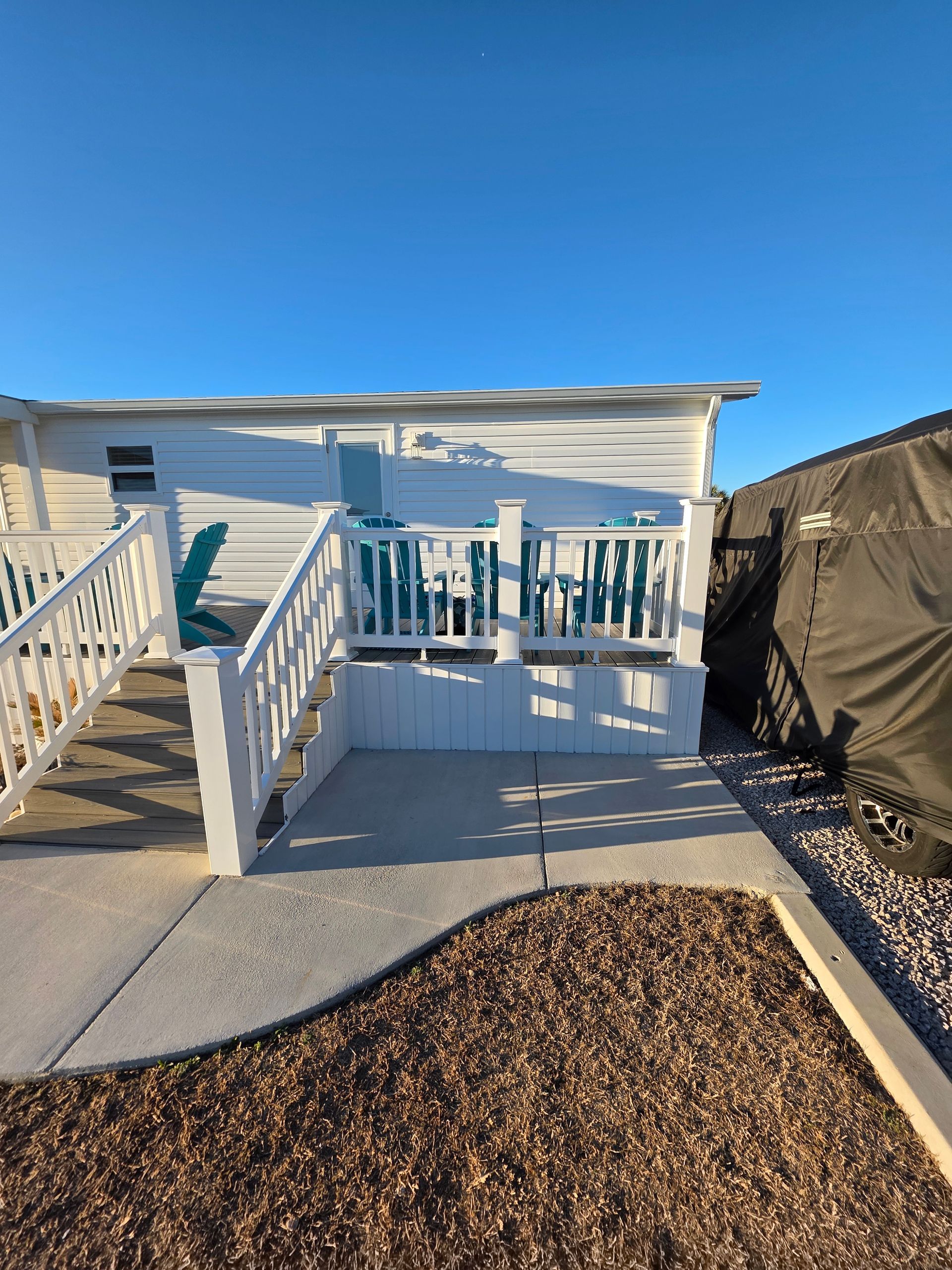 White deck and staircase with turquoise chairs outside a mobile home, next to a covered area on a sunny day.