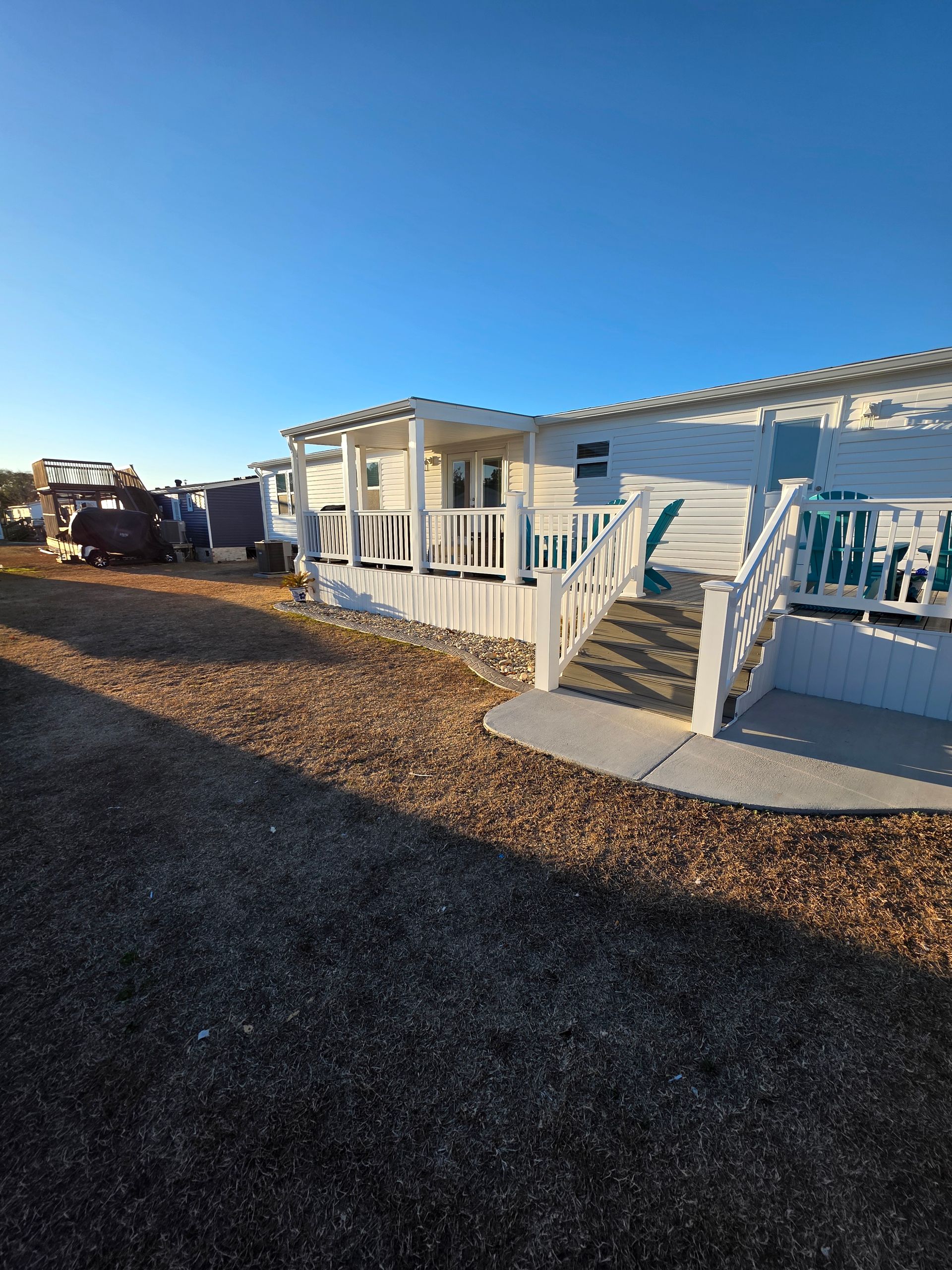 White mobile home with a white porch and ramp, set in a sunny, gravel lot under a clear blue sky.
