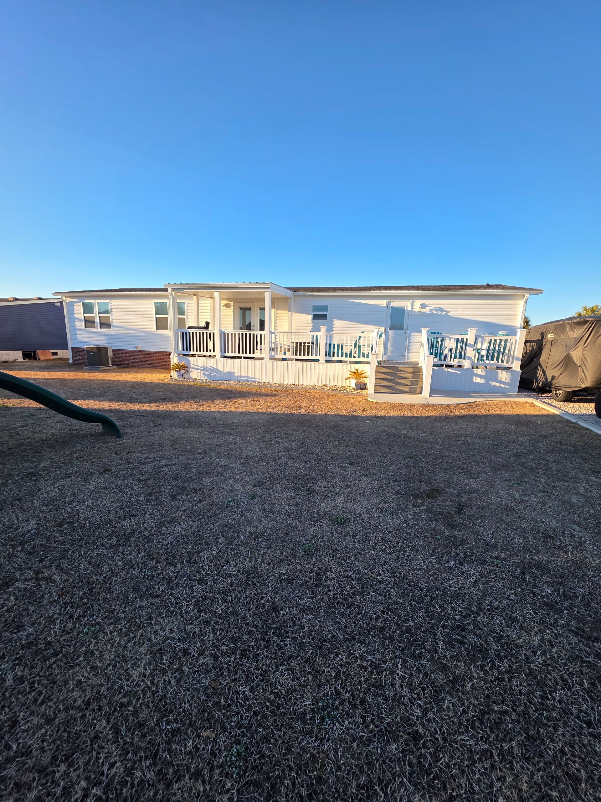 A light-colored mobile home with a white porch sits on a gravel lot under a clear blue sky.