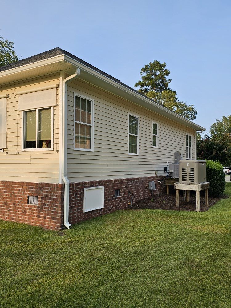 Side view of a light-colored, vinyl-sided home with a brick foundation, featuring an outdoor HVAC unit on a raised stand.