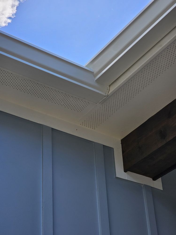Low-angle view of a blue house corner where the roof soffit meets a dark wood beam against a blue sky.