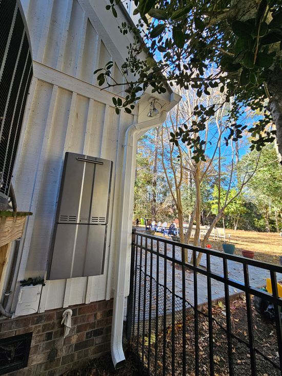 A vertical tankless water heater mounted on the white siding of a house, next to a white downspout and a black fence.