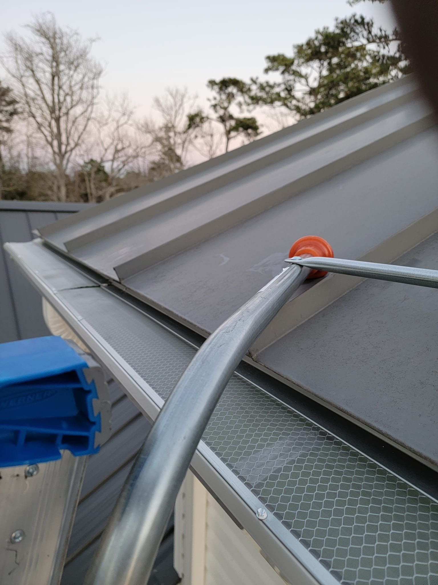 A metal conduit pipe with an orange fitting is installed along the edge of a metal roof, resting over a gutter.