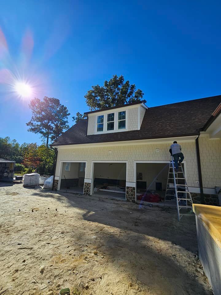 A worker on a ladder performs construction on the exterior of a garage with three bays and a dormer under a bright sun.