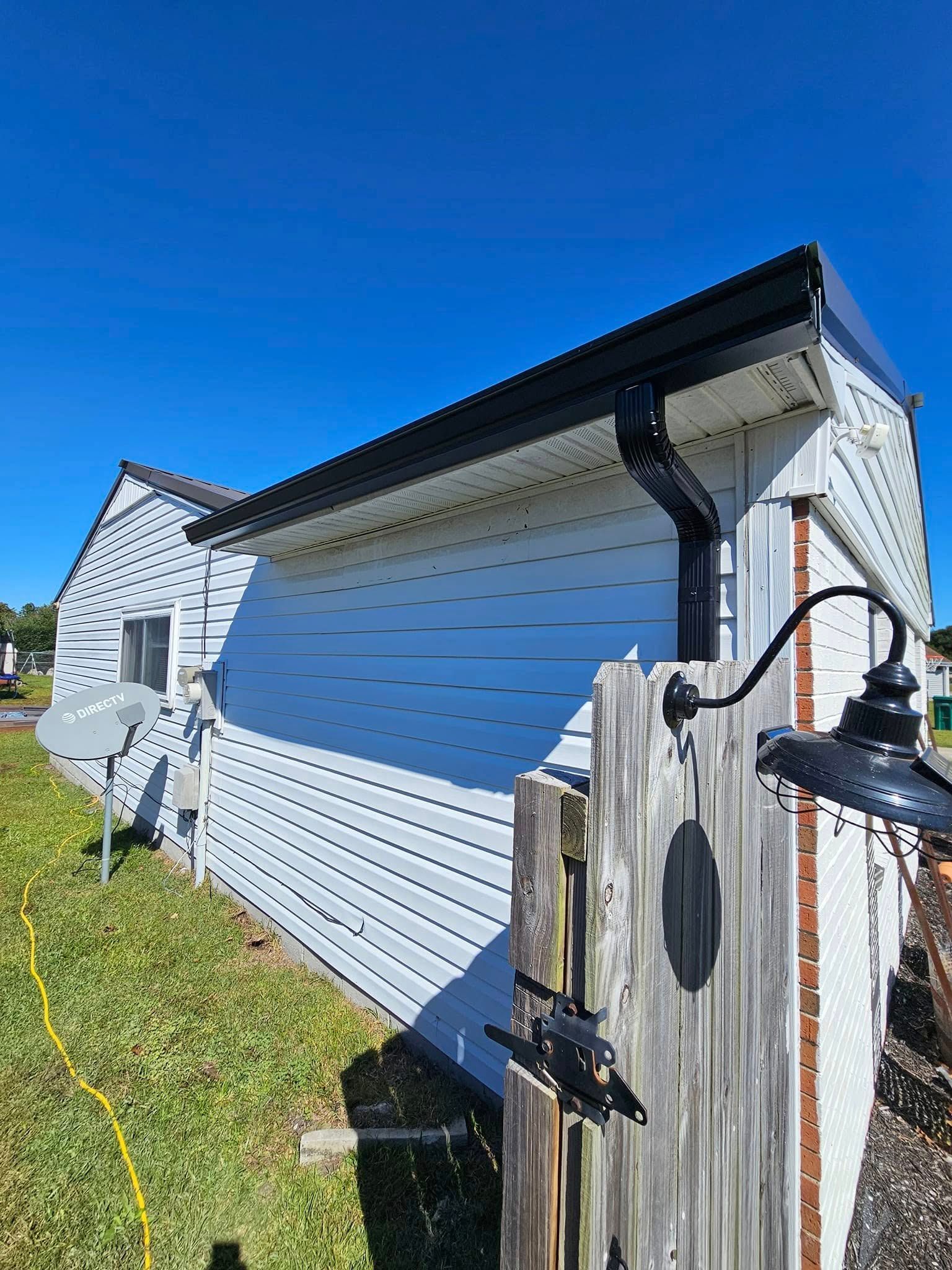 A side view of a light blue vinyl-sided house under a bright blue sky, featuring a black gutter and a wooden gate nearby.