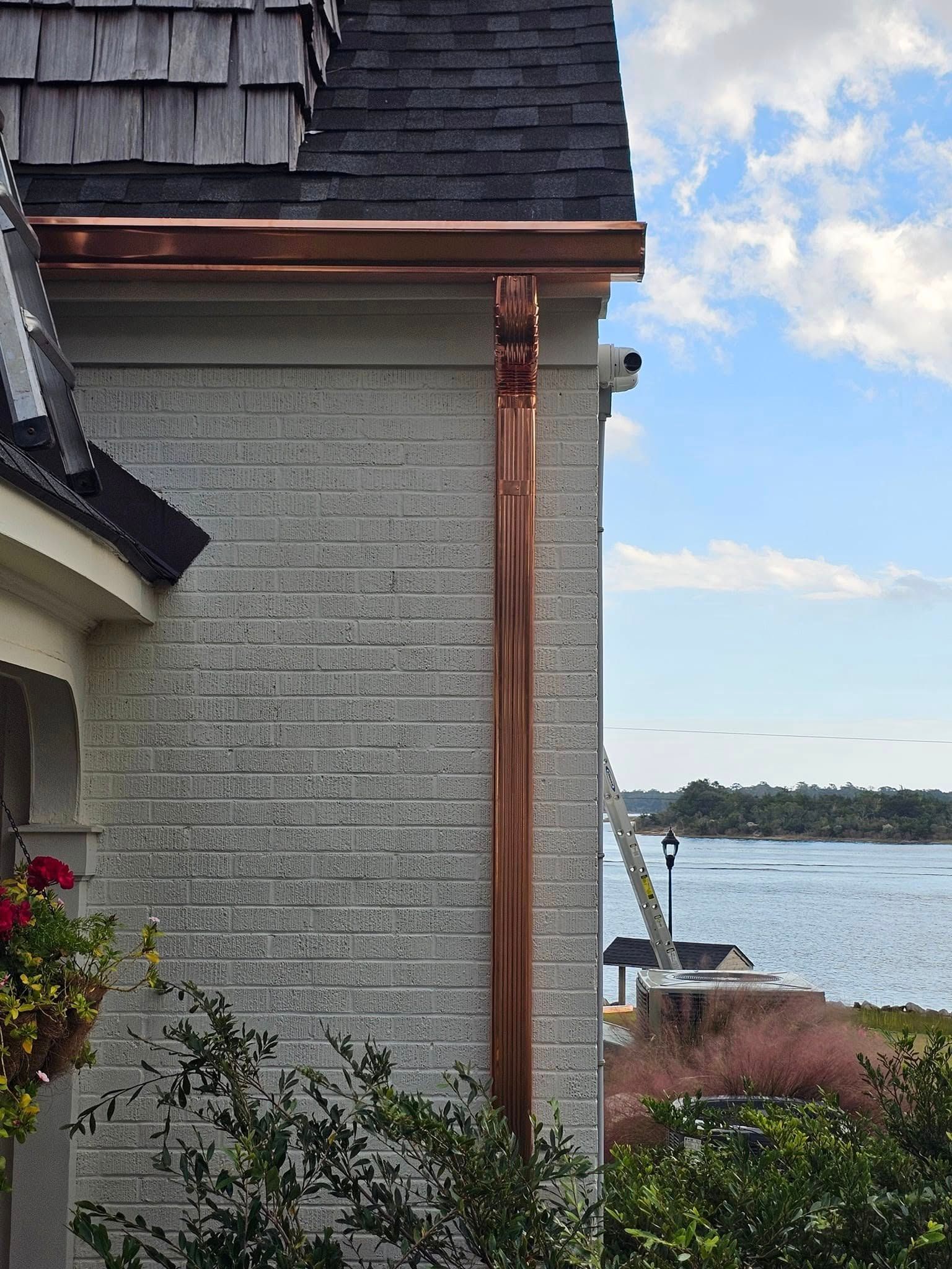 Copper gutter and downspout installed on the corner of a light-colored brick house overlooking a coastal waterway.