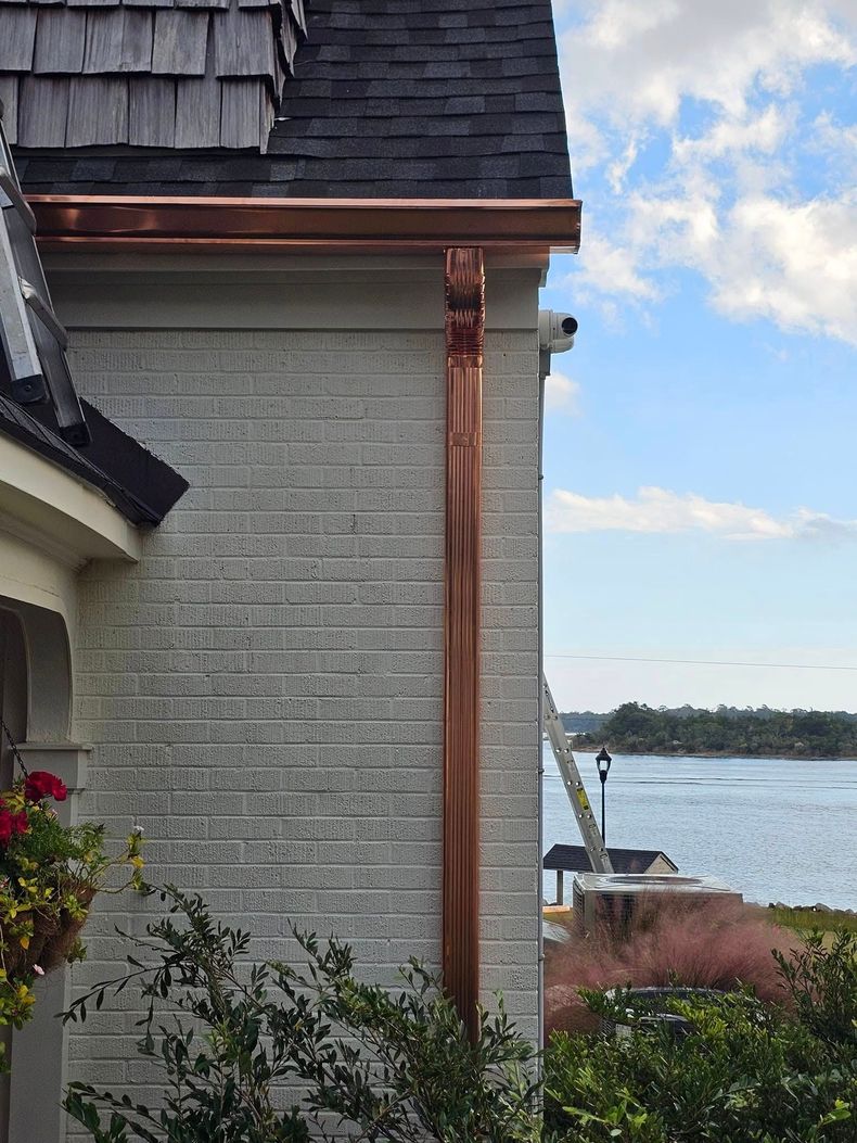 Copper gutter and downspout installed on the corner of a light brick house overlooking a coastal water view.