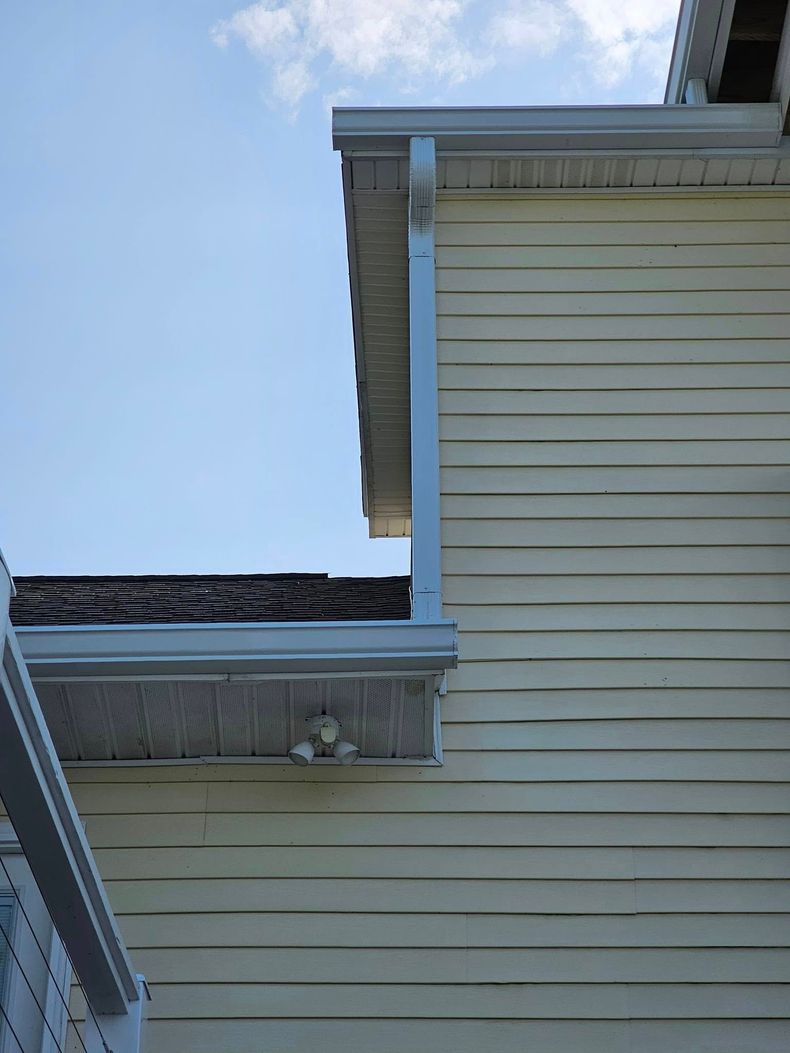 Exterior corner of a yellow-sided house with white trim, a dark shingled roof overhang, and a metal gutter downspout.