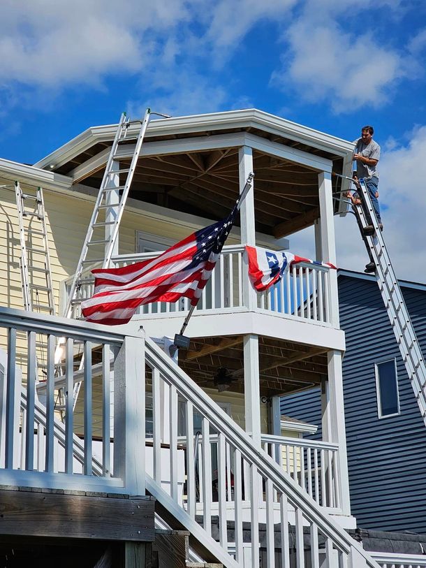 A worker stands on a ladder against the second story of a house, which features a porch and an American flag.