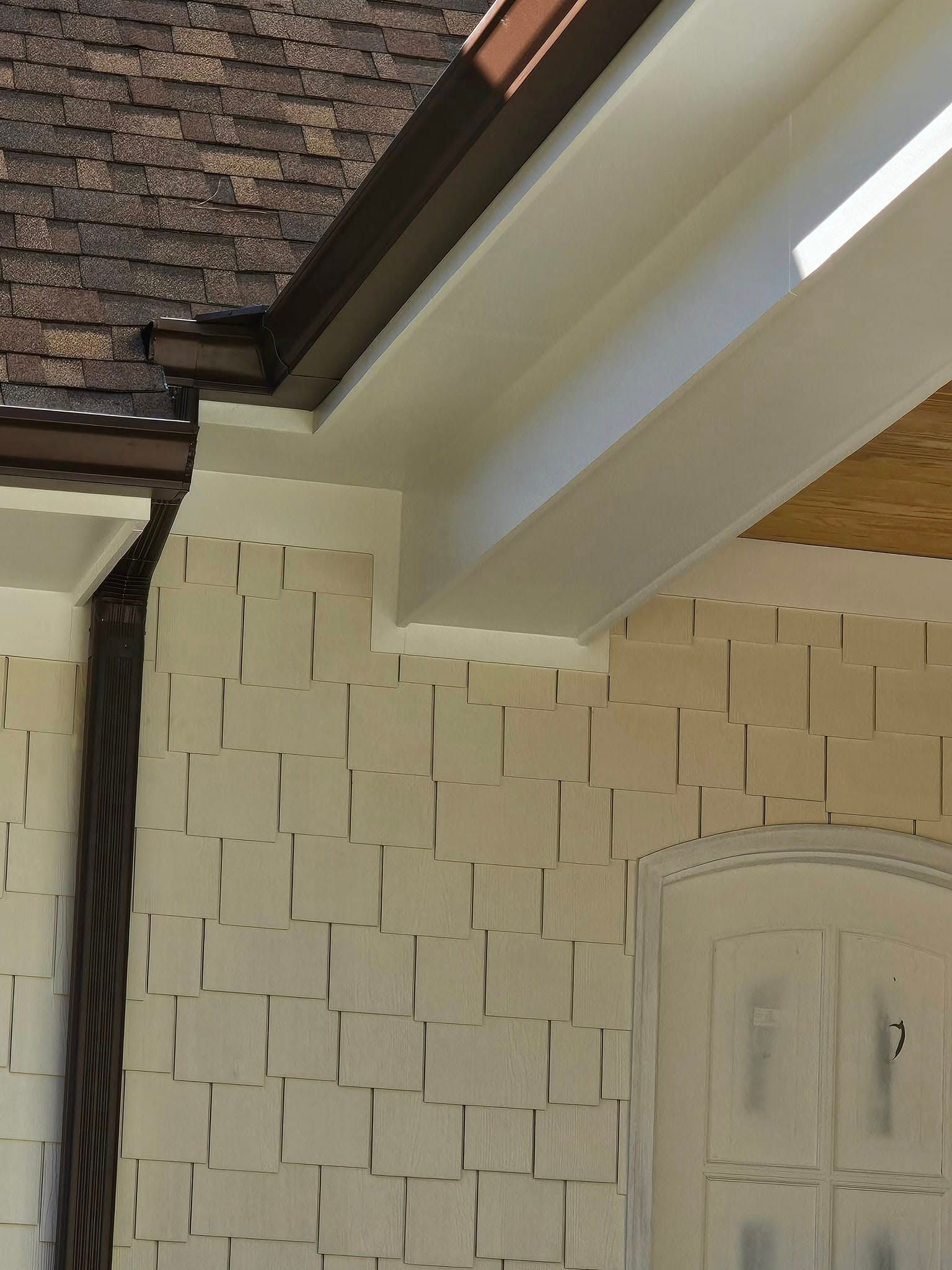 Exterior corner of a house showing beige shingle siding, a brown gutter, and a white soffit near an arched doorway.