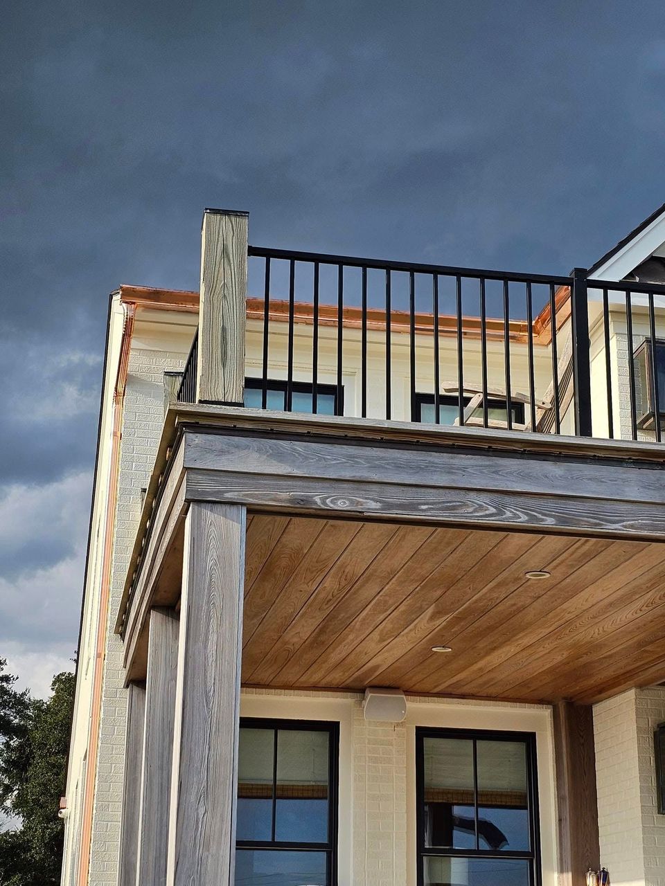 An exterior view of a balcony featuring natural wood beams and pillars above two windows under a cloudy sky.