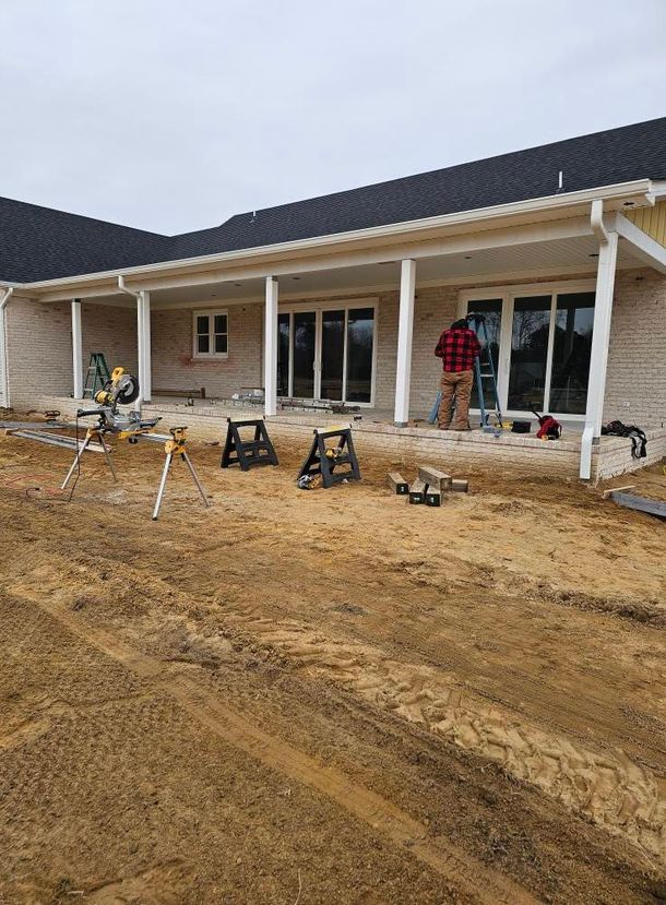 A construction worker installing trim on a covered patio with a brick exterior and a dirt ground in front of the house.