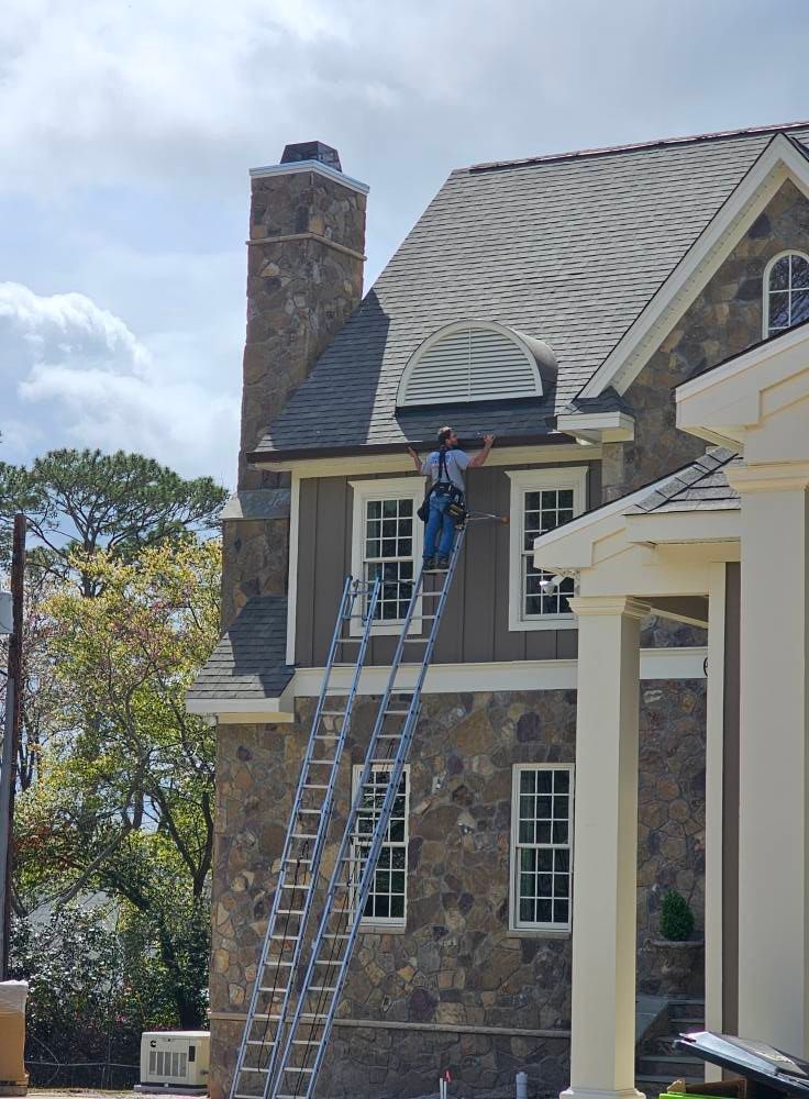 A worker in a harness stands on an extension ladder to perform roof maintenance on a two-story stone house.