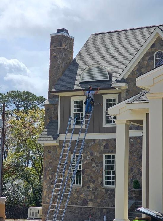 A worker stands on a tall ladder, performing exterior maintenance on the second-story roof of a stone and siding house.