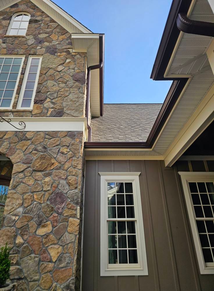 Low-angle view of a house exterior featuring a stone wall, tan vertical siding, and dark gutters against a clear blue sky.