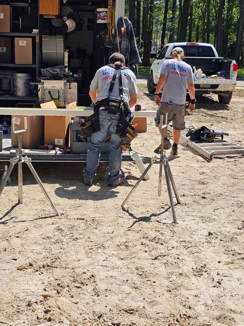 Two workers wearing grey shirts and tool belts work outside near a trailer, a white truck, and metal equipment stands.
