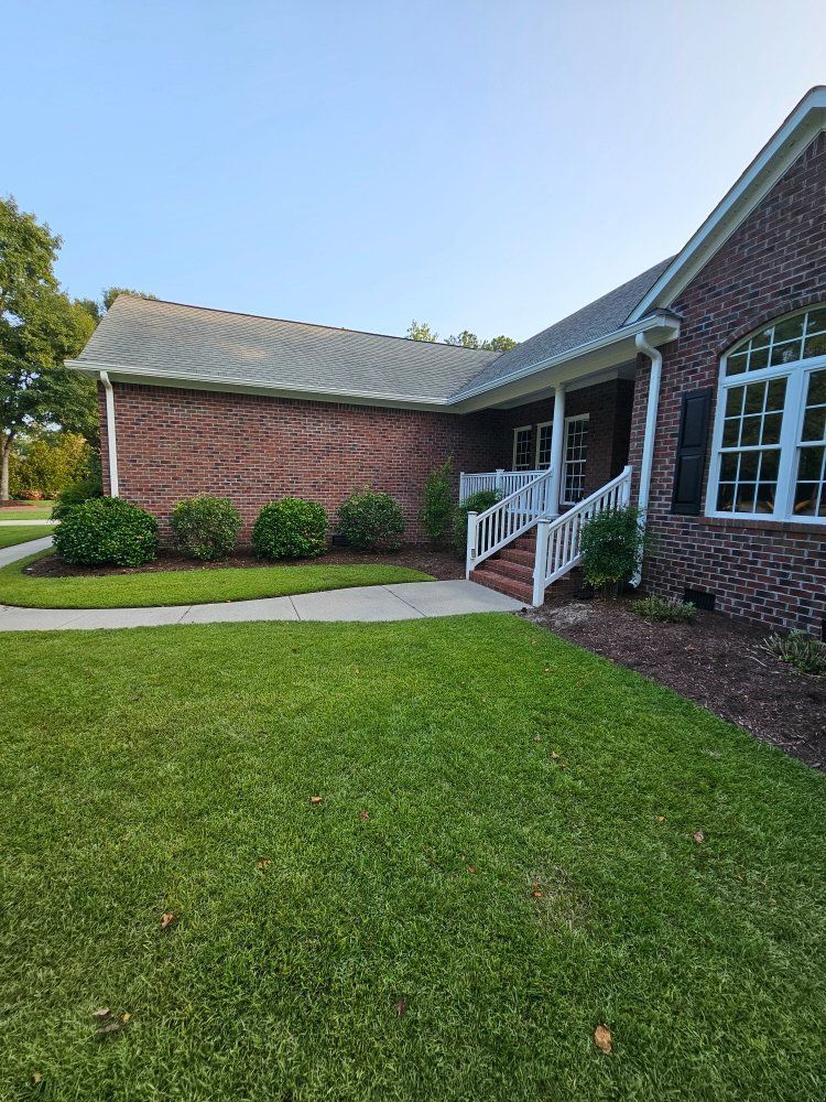 A red brick house with a white front porch, grey shingled roof, and green lawn and shrubs under a clear blue sky.