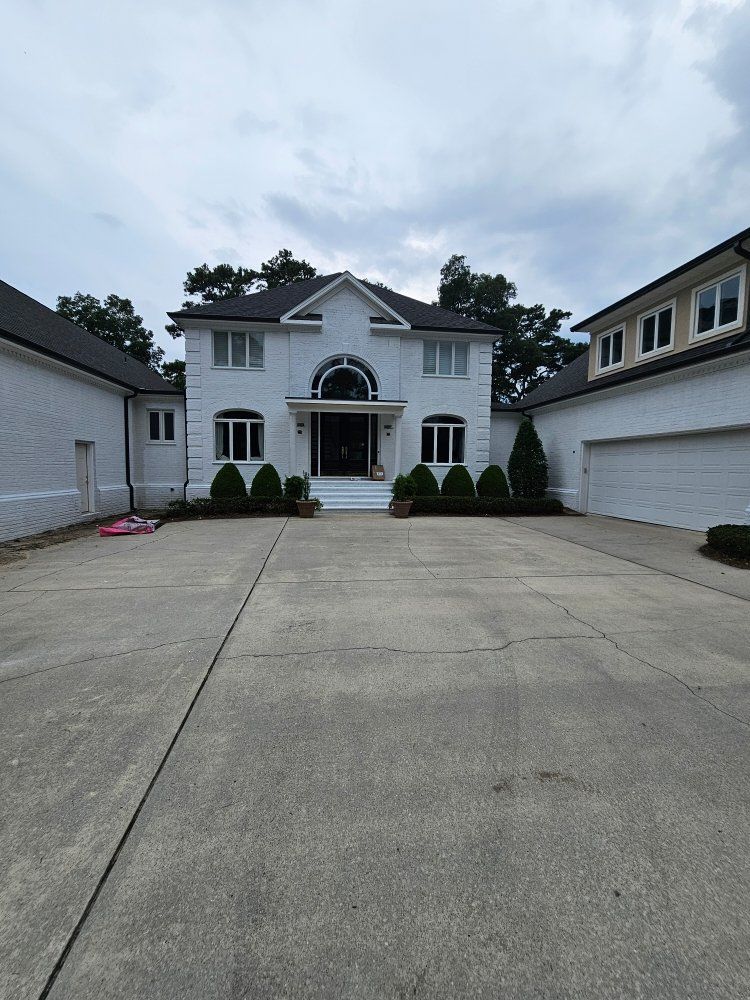 A white, two-story house with a large paved driveway, dark roof, and symmetrical windows under a cloudy sky.