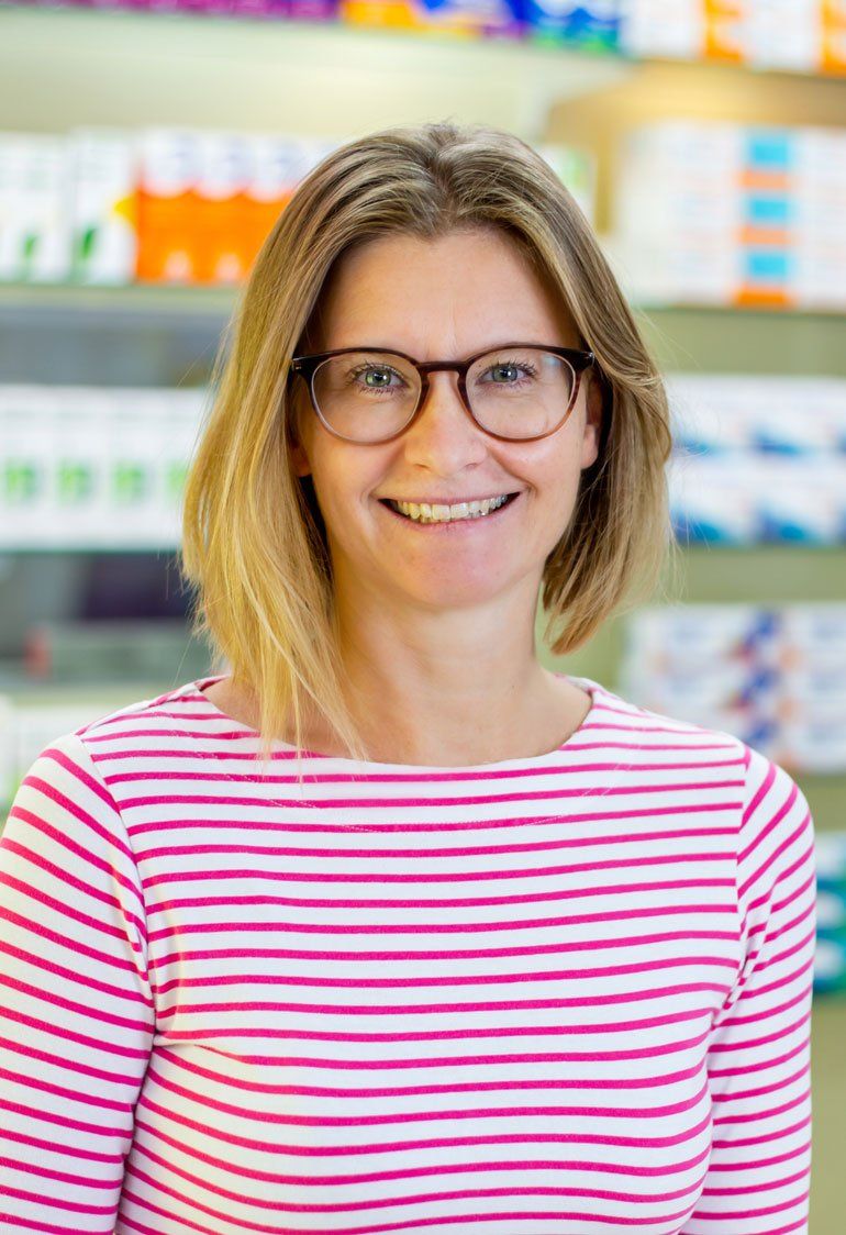 Eine Frau mit Brille und rosa gestreiftem Hemd lächelt in einer Apotheke.