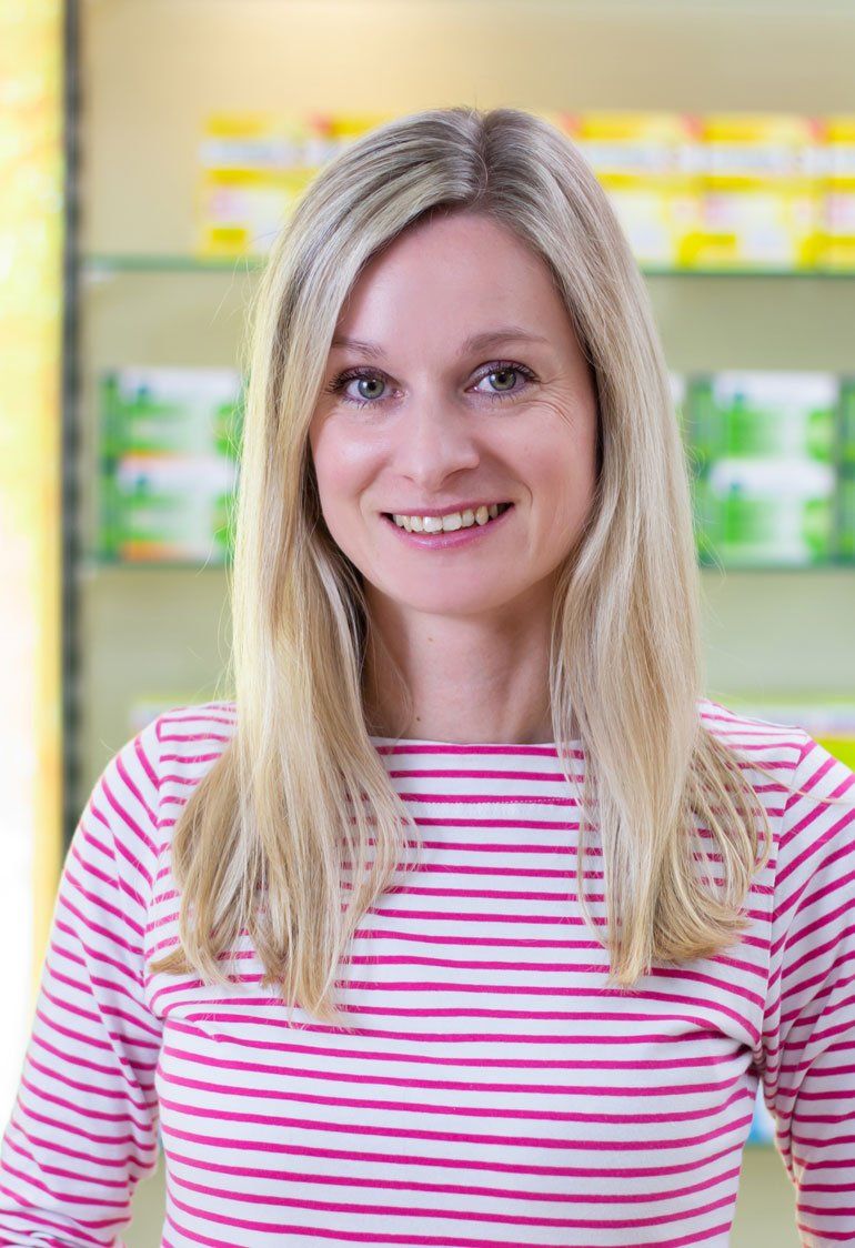 Eine Frau in einem rosa-weiß gestreiften Hemd lächelt in einer Apotheke.