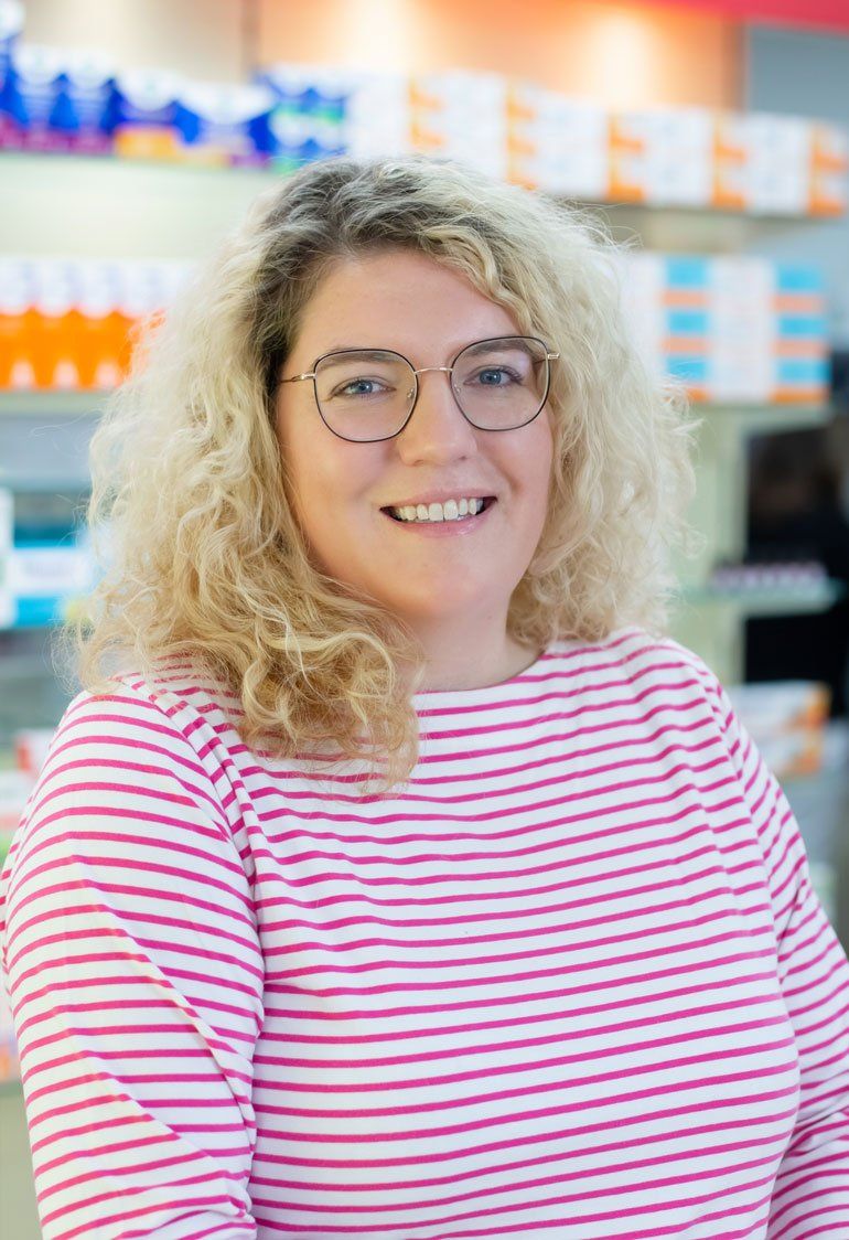 Eine Frau mit Brille und rosa-weiß gestreiftem Hemd lächelt in einer Apotheke.