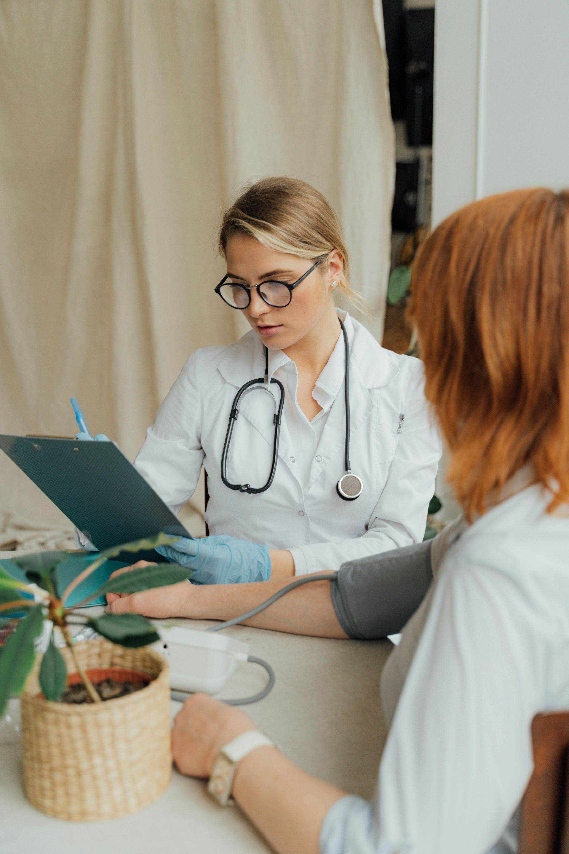 Doctor taking patient's blood pressure, writing on a clipboard. The doctor wears glasses and a stethoscope.