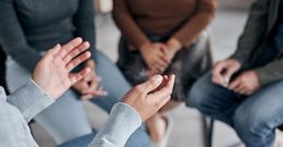 Person gesturing with hands, seated in a circle with others; possibly a support group.