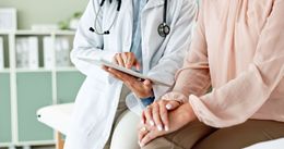 Doctor in white coat reviewing tablet with patient, hands clasped. Medical setting.
