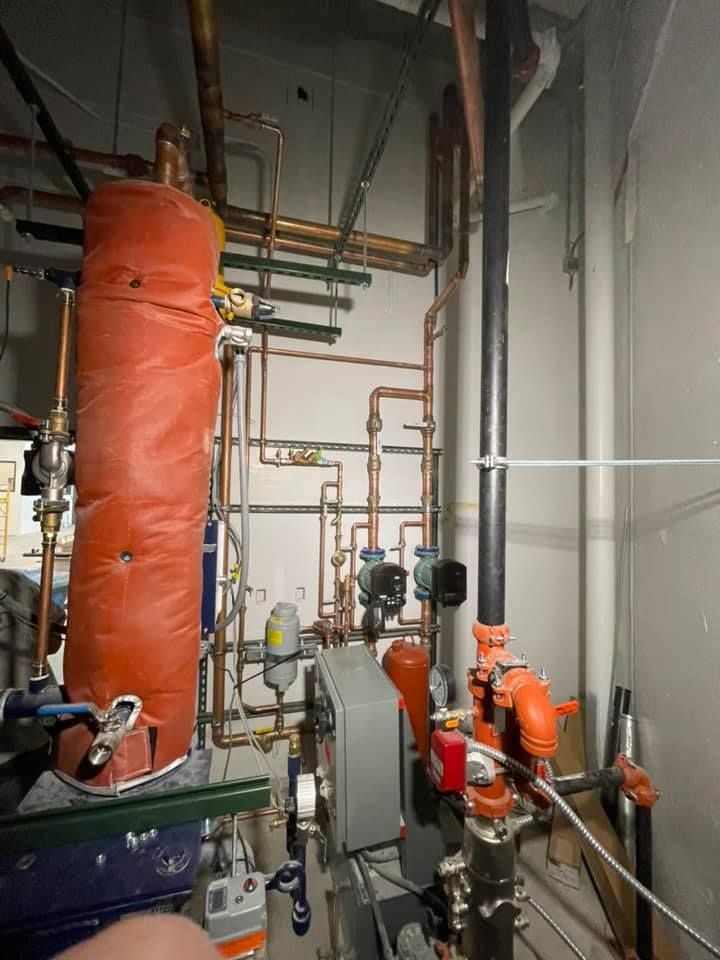 Industrial mechanical room with pipes, valves, tanks, and orange insulated equipment along a narrow corridor
